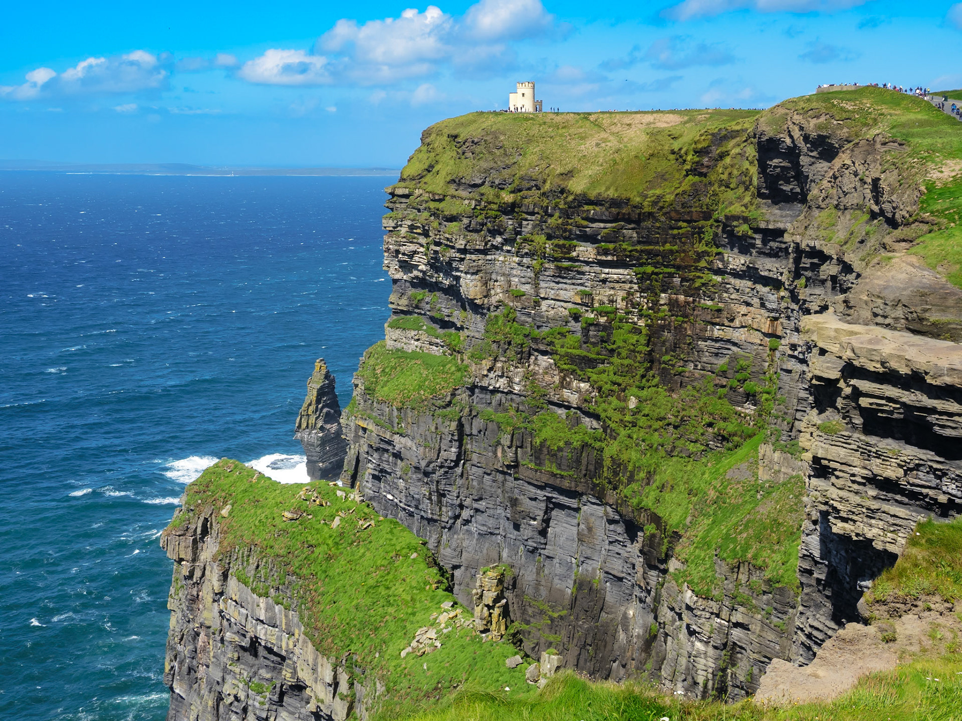 Das Bild zeigt die berühmten Cliffs of Moher an der irischen Westküste. Im Vordergrund ragen die steilen, mit Gras bewachsenen Felsen dramatisch aus dem Atlantik. Auf dem Plateau steht der O'Brien's Tower, ein kleiner historischer Aussichtsturm. Das Meer ist tiefblau und leicht bewegt, der Himmel ist größtenteils blau mit einigen weißen Wolken. Die Perspektive ist leicht erhöht und vermittelt einen weiten Blick entlang der Küste. Die Stimmung ist klar, frisch und lebendig, typisch für einen sonnigen Tag an der irischen Küste.