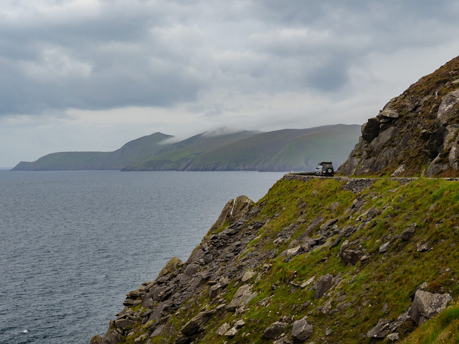 Das Bild zeigt eine dramatische Küstenlandschaft mit einer schmalen, kurvigen Straße, die sich entlang einer steilen Klippe schlängelt. Ein Geländewagen fährt auf dieser Straße. Das Meer erstreckt sich im Hintergrund, und im fernen Hintergrund sind grüne Hügel oder Inseln zu sehen. Der Himmel ist bedeckt mit grauen Wolken, was eine düstere und atmosphärische Stimmung erzeugt. Die Farben sind gedämpft und hauptsächlich in Grün-, Grau- und Blautönen gehalten. Die Perspektive ist leicht erhöht, was einen guten Überblick über die Landschaft ermöglicht.