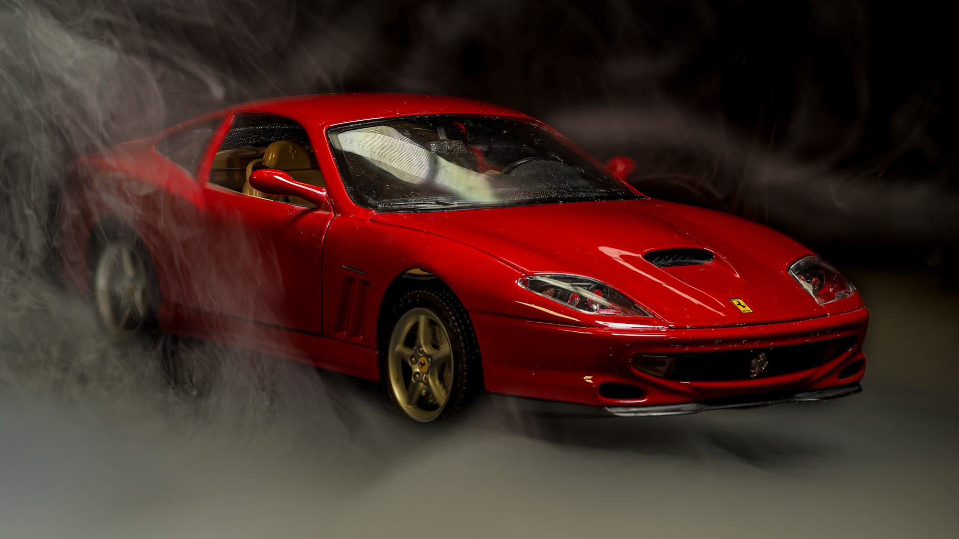 Das Bild zeigt ein rotes Modellauto eines Sportwagens, das von Rauch umgeben ist. Die Aufnahme ist nah und fokussiert auf das Auto, der Hintergrund ist dunkel und unscharf. Die Lichtführung hebt die glänzende Oberfläche des Autos hervor und erzeugt eine dramatische, mystische Stimmung.