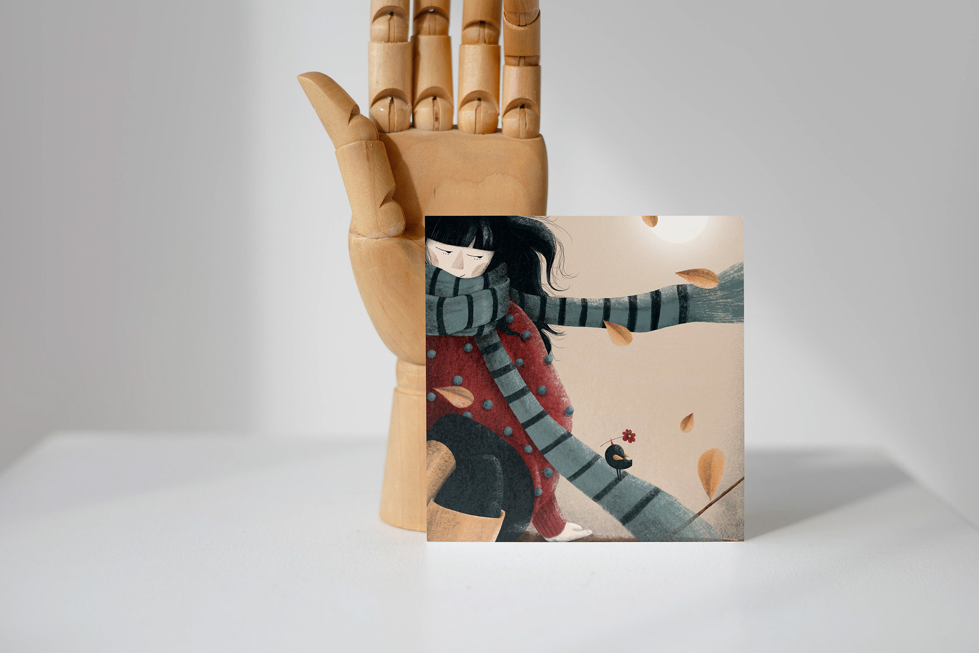 Small illustrated art print of 'You and Me, Valerie' by Darka White Art, propped against a wooden hand model, depicting a girl with a flowing scarf and a bird, surrounded by autumn leaves. A unique custom print for art enthusiasts.