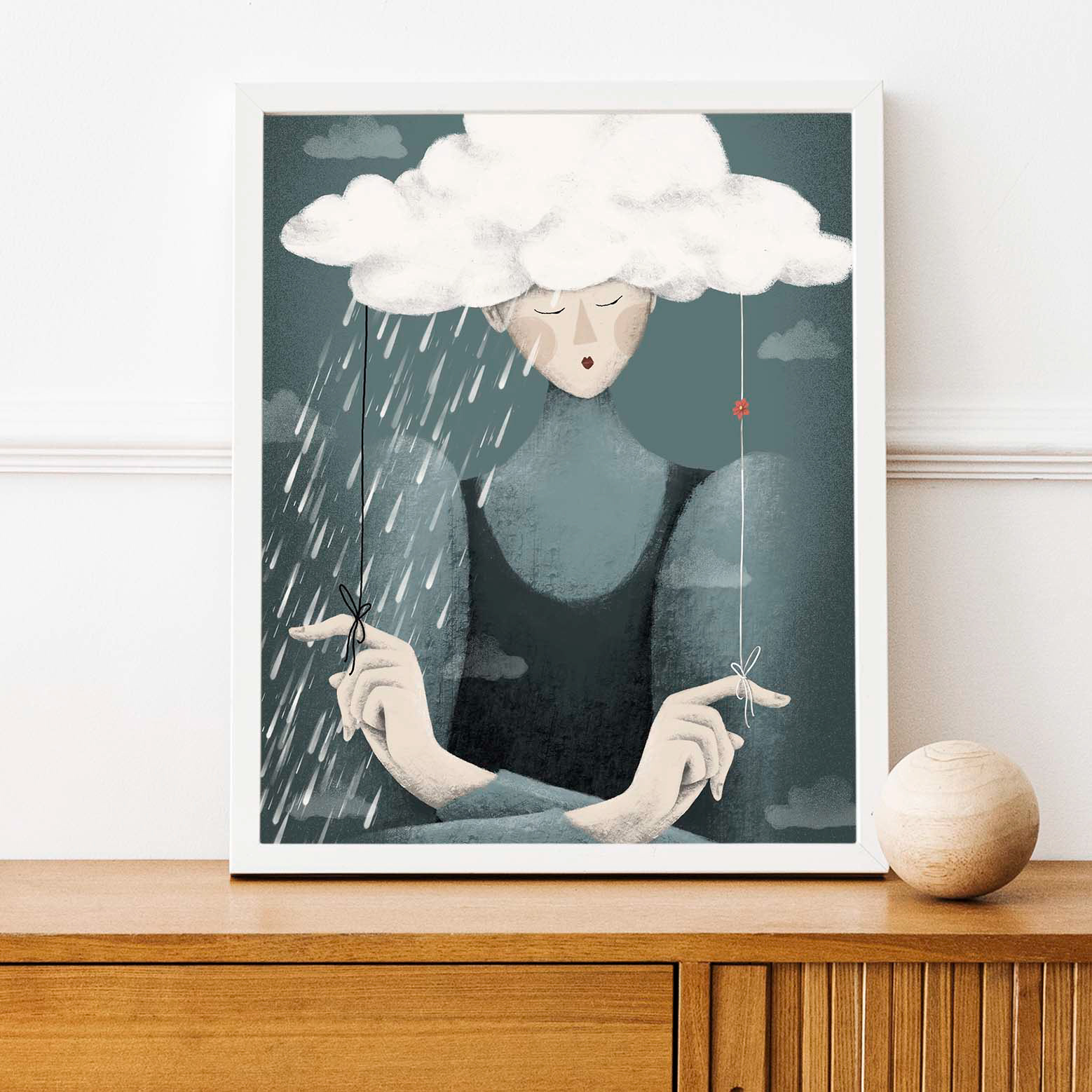 Framed 'Moody Mandy' art print by Darka White Art, featuring a pensive character with a cloud over her head, symbolizing introspection and rain, displayed on a wooden sideboard for home decor.