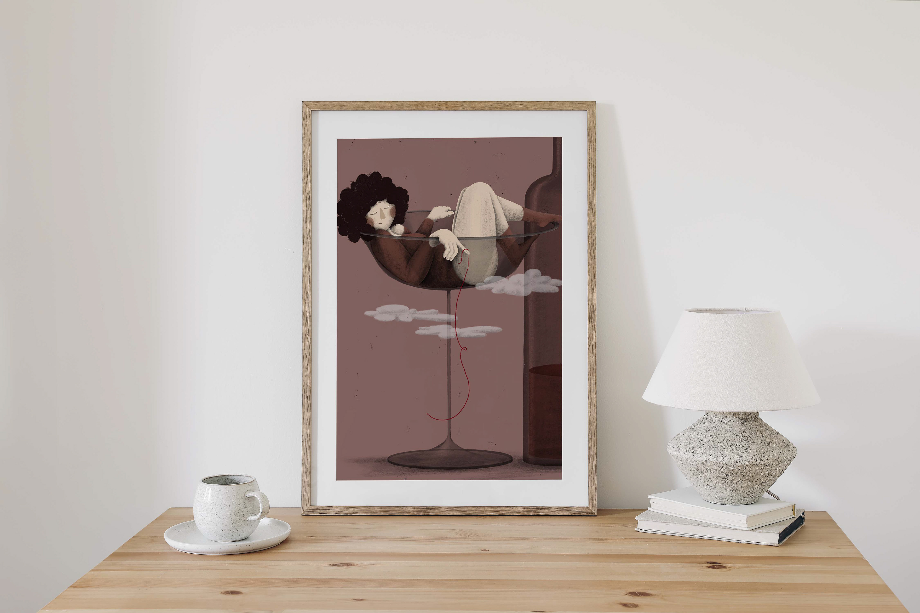 A framed illustration by Darka White Art depicts a peaceful woman lounging in a wine glass, accompanied by a bottle and clouds, and placed on a minimalist wooden table with books and a lamp.