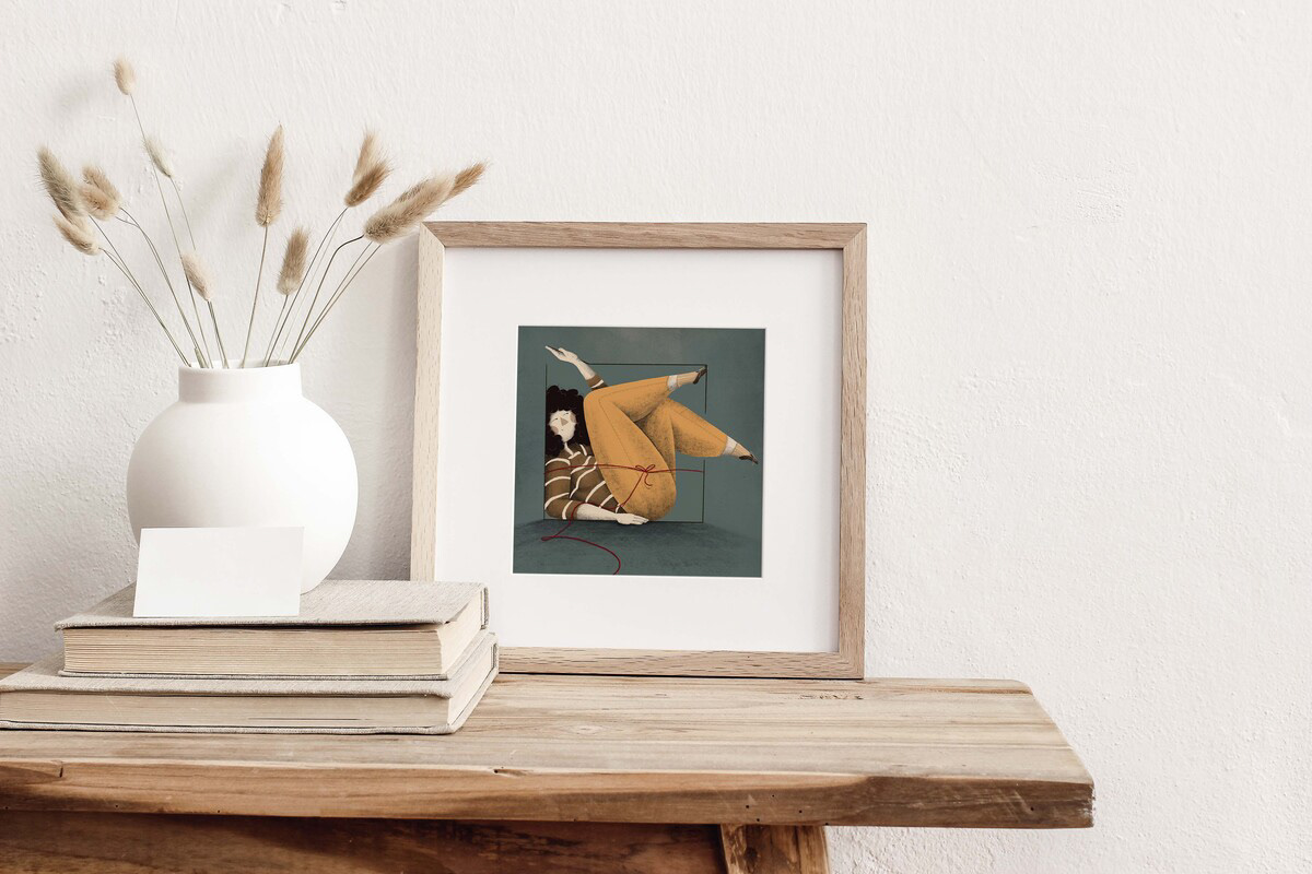 Unique framed art print of a woman in yellow pants and striped shirt, tied with red string, displayed on a rustic wooden table next to a white vase and books - Darka White Art Illustration.