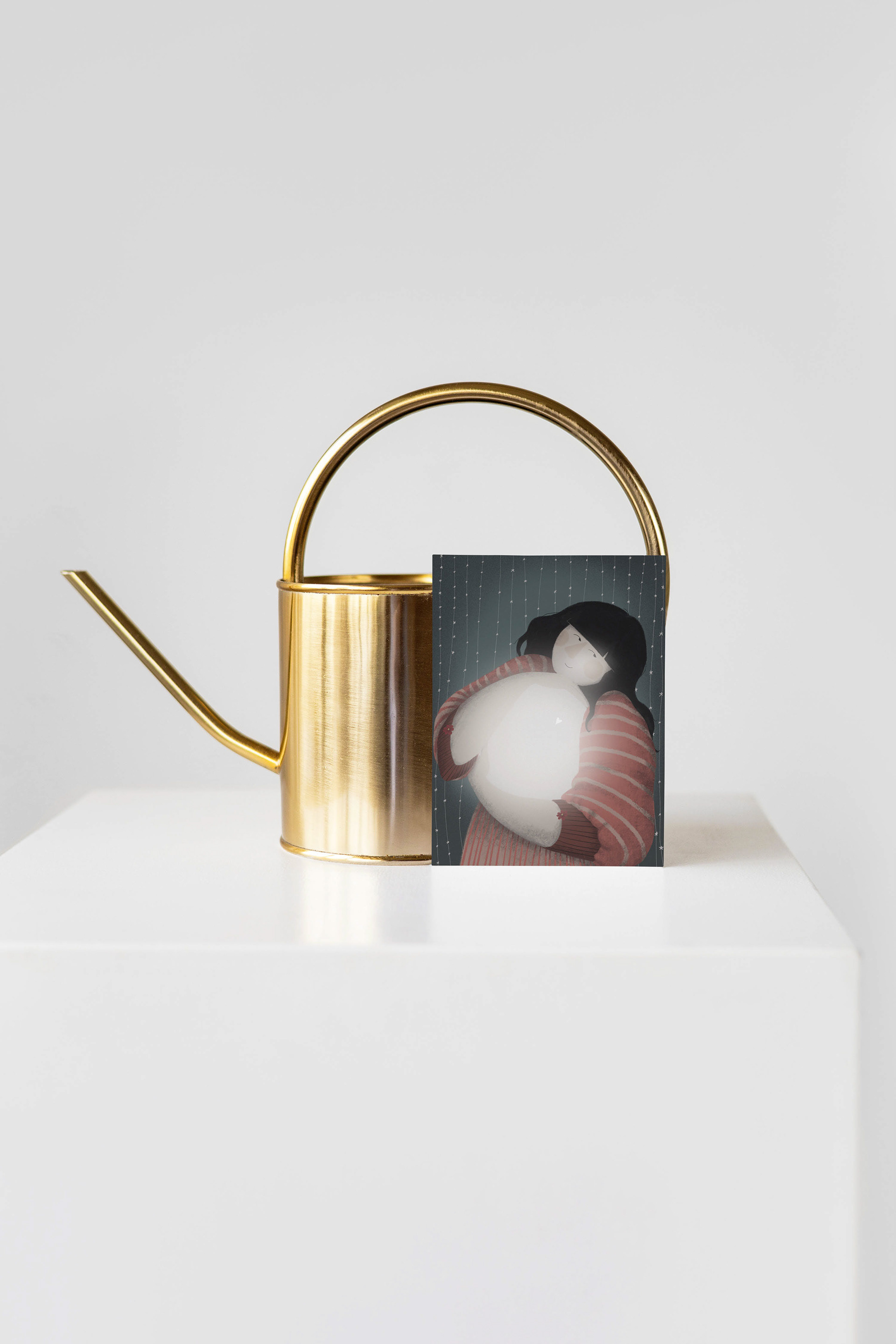The Way to Self' art print by Darka White Art, placed beside a modern gold watering can on a minimalist white surface, depicting a person in a striped sweater hugging a ball of light with a backdrop of delicate, starry strings, evoking self-love and warmth.