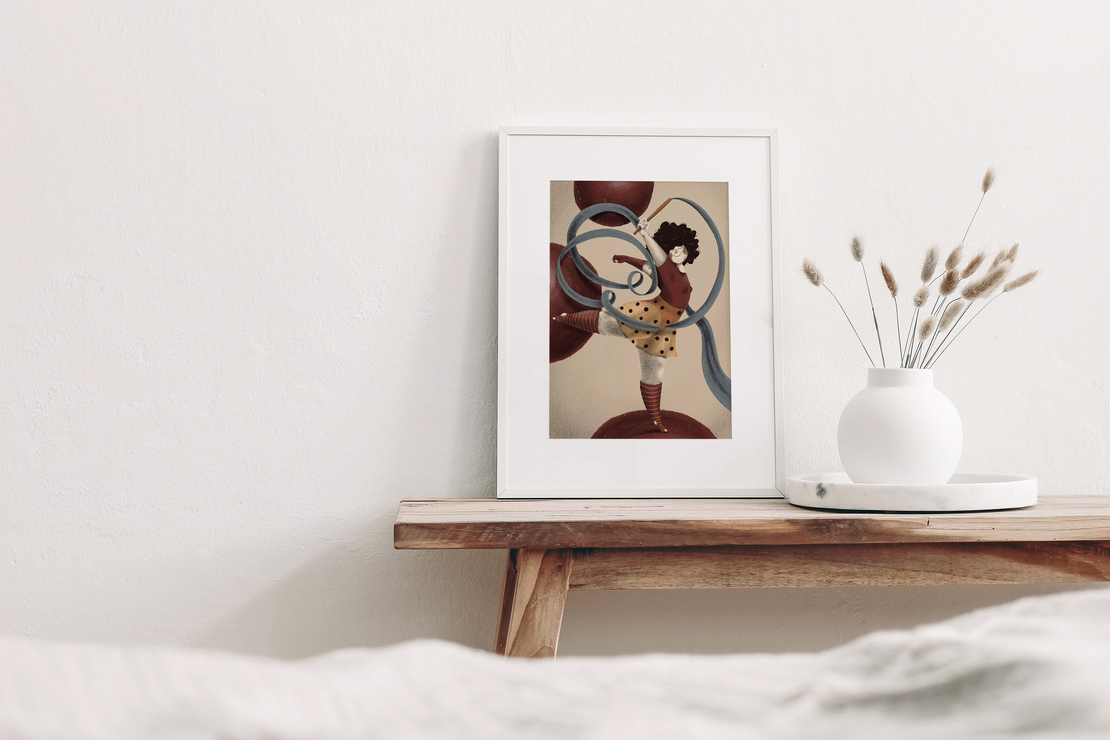 Framed illustration of a whimsical dancer twirling a ribbon, placed on a wooden bench against a white wall. The artwork, featuring a curly-haired dancer in a polka-dotted skirt and red top, is accompanied by a white vase holding dried grasses, creating a minimalist and serene interior setting. Illustration by Darka White Art