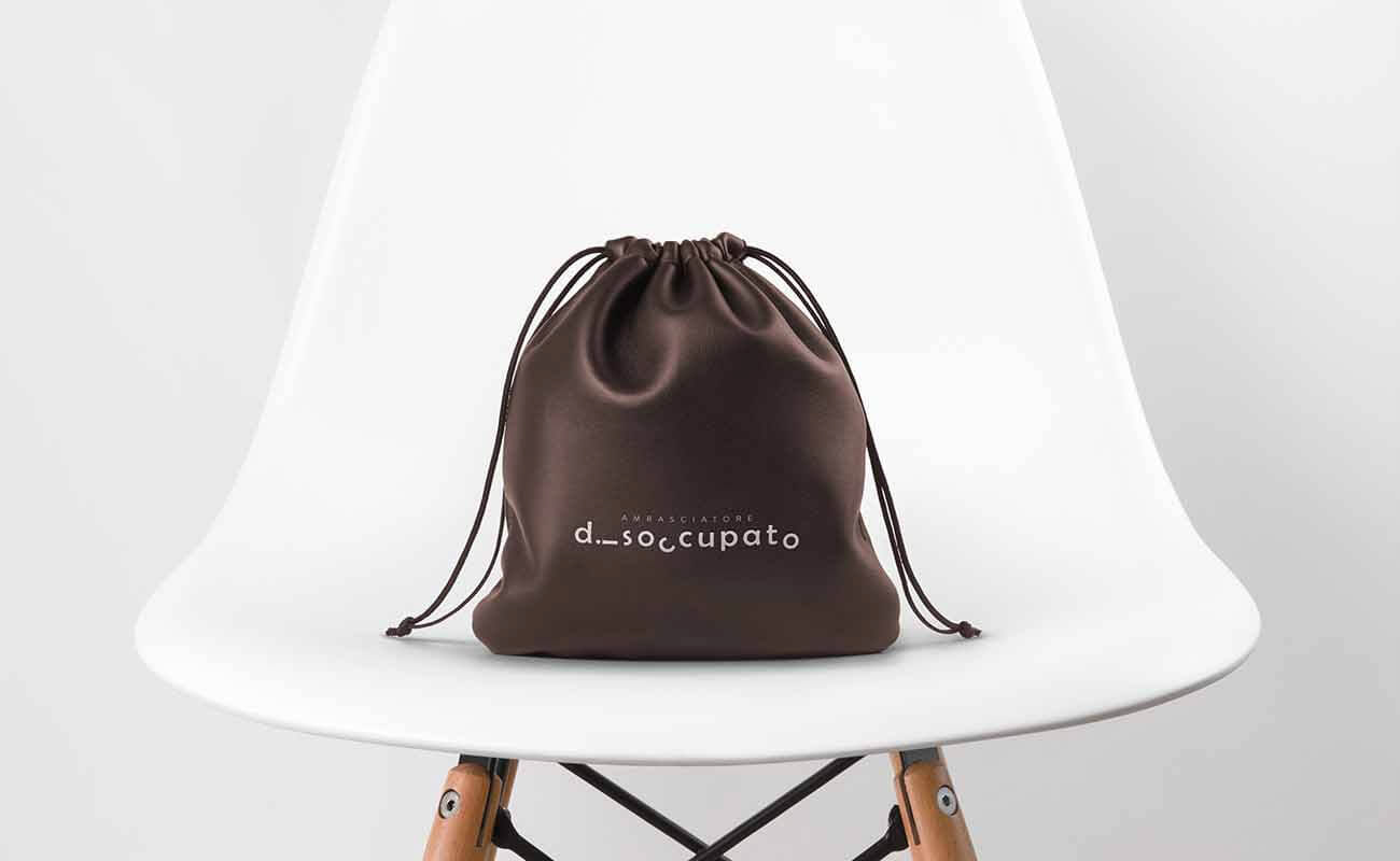 Logo of 'Ambasciatore Disoccupato' designed by graphic designer Darka White, displayed on a brown backpack placed on a chair, reflecting the client's adventurous spirit with dynamic lettering.