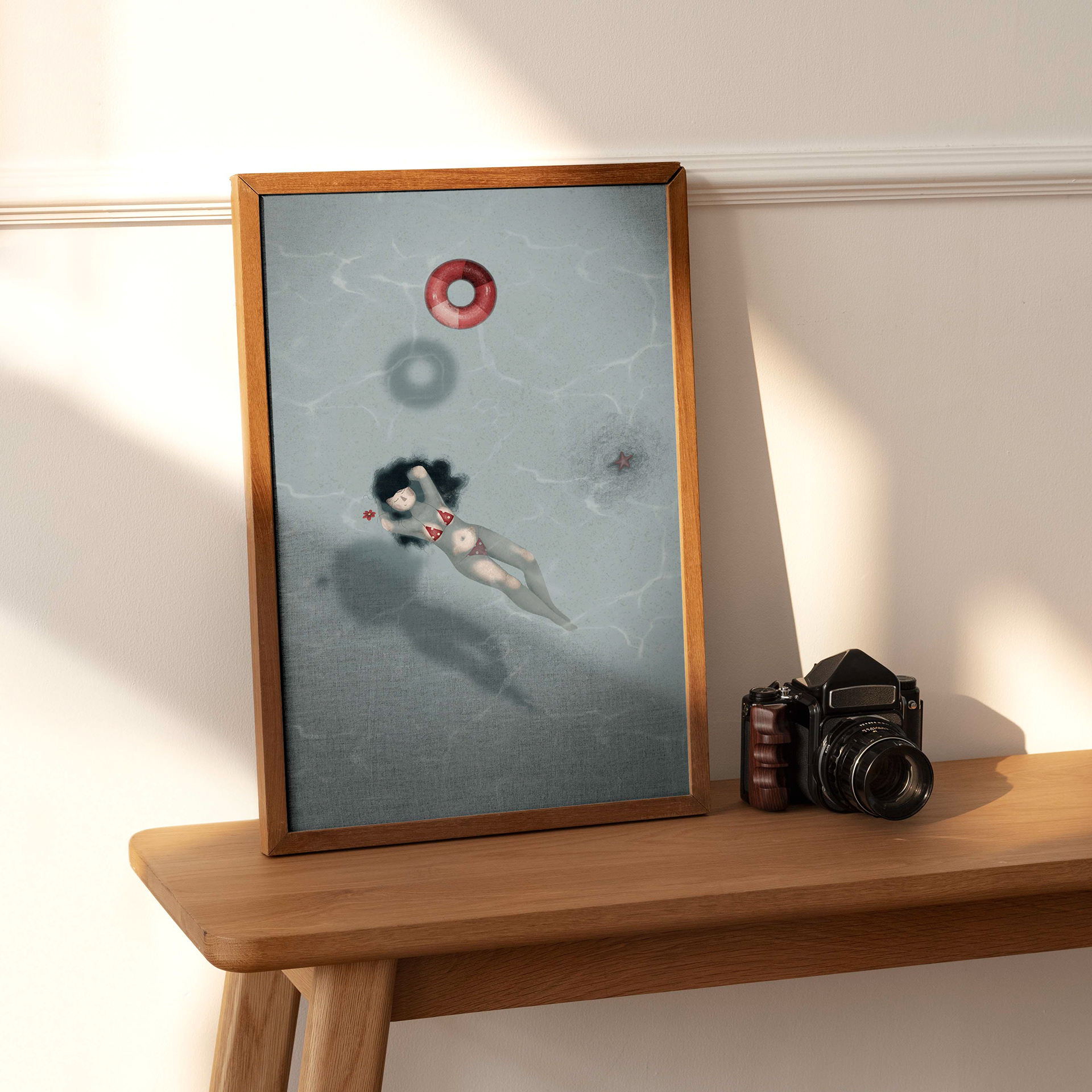 Digital illustration by Darka White Art displayed in a wooden frame on a light-colored table next to a vintage camera. The artwork features a woman in a red bikini floating peacefully in a clear pool, with a life preserver, red flower, and starfish, capturing a serene and tranquil moment.