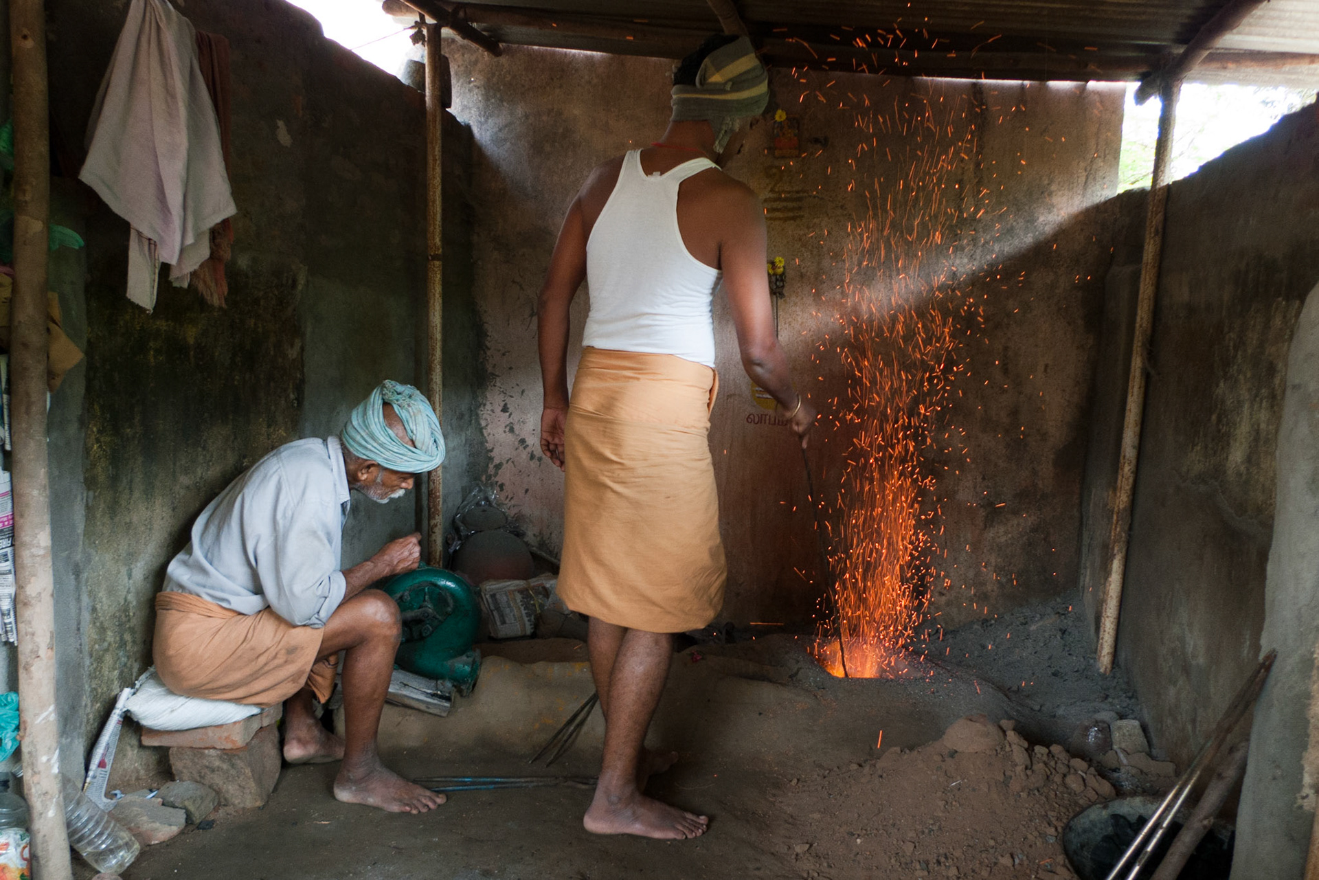 Thanjavur: Bronze casting. The older man is hand winding a fan connected by a tunnel to the furnace. (Both wearing appropriate  gear.)