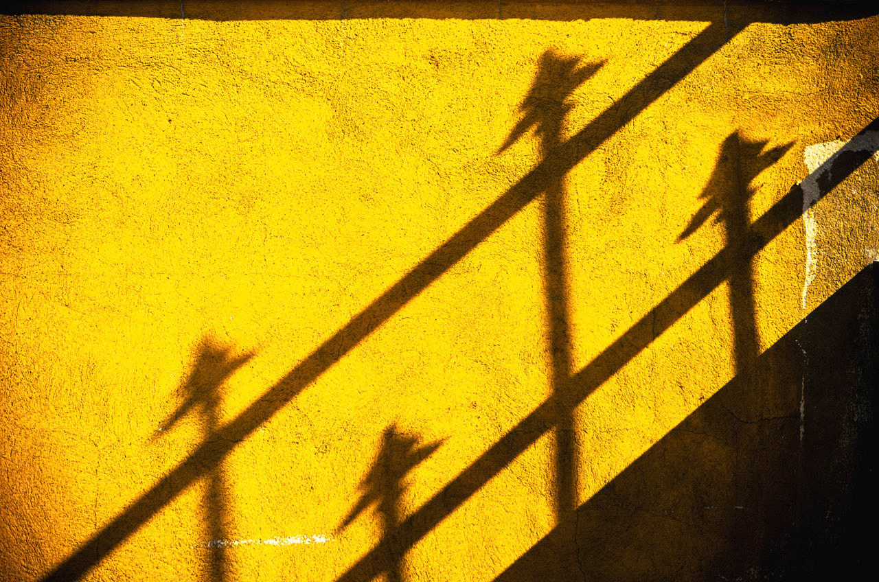 Fence detail shadows of four spears on an old rough yellow wall.