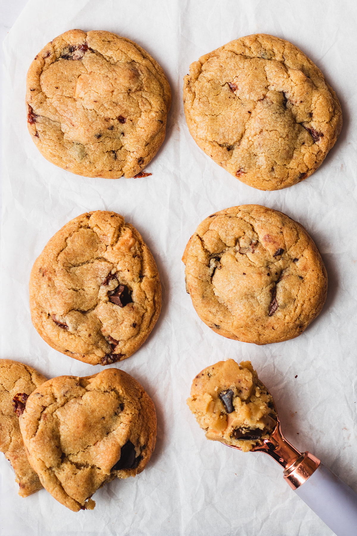Five chocolate chip cookies arranged on white paper with one broken open