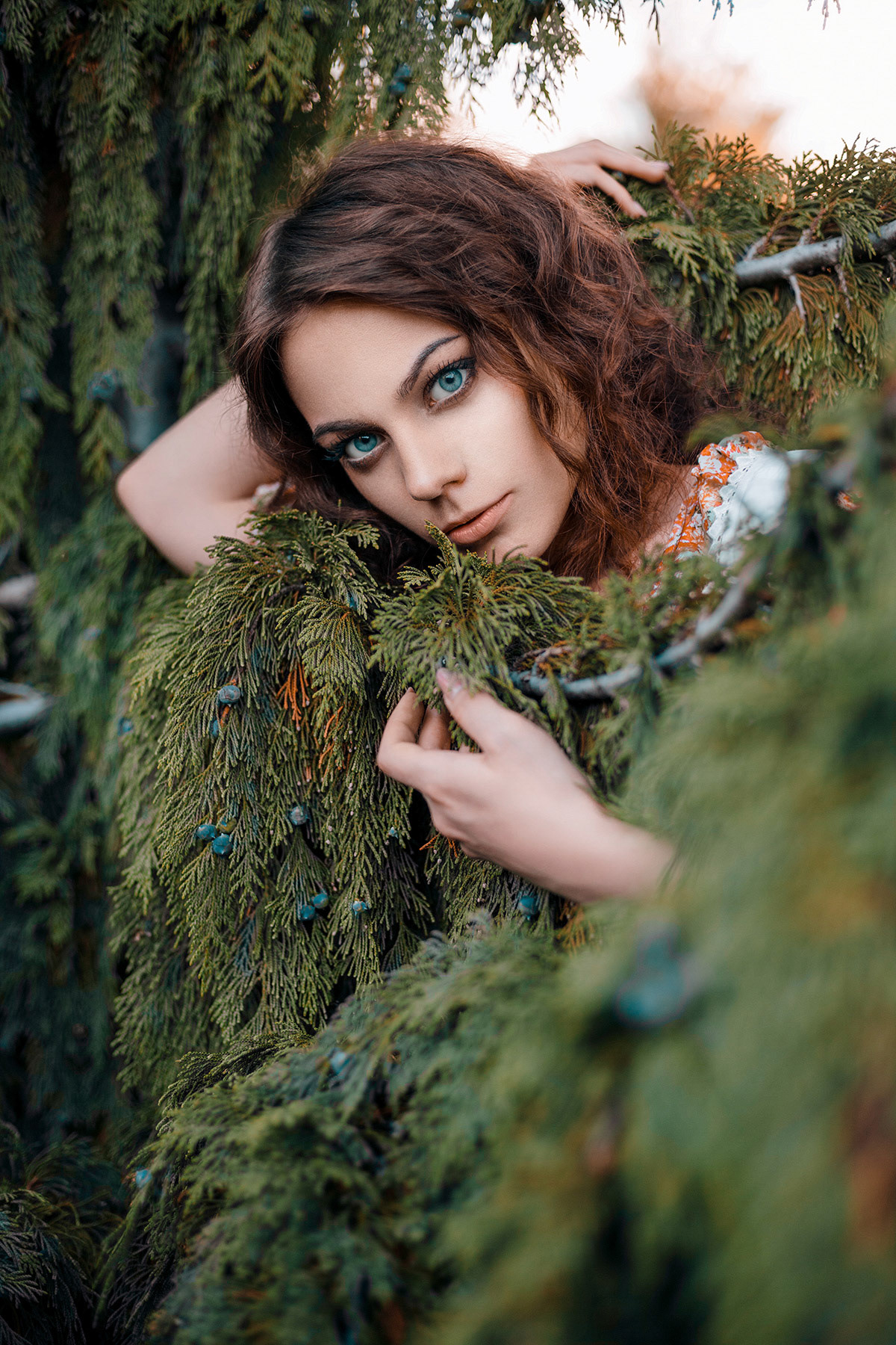 Close-up summer portrait of a beautiful curly haired girl in garden. Pretty young lady hugs evergreen plant. Perfectly retouched.