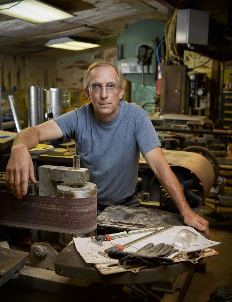 A man leaning on one of his mid-century industrial woodworking machines that is housed in his barn woodworking shop.