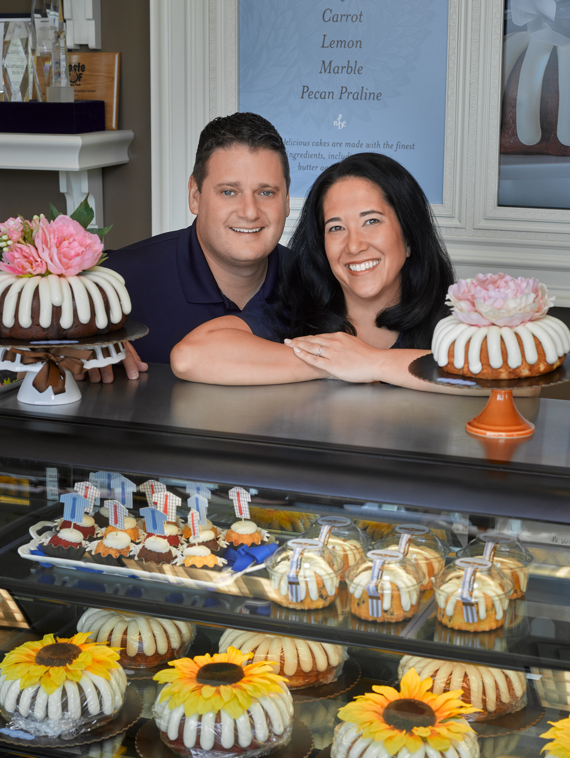 Owners of a specialty bakery pose behind a colorful display of signature bundt cakes and seasonal treats created for their retail storefront and marketing materials.