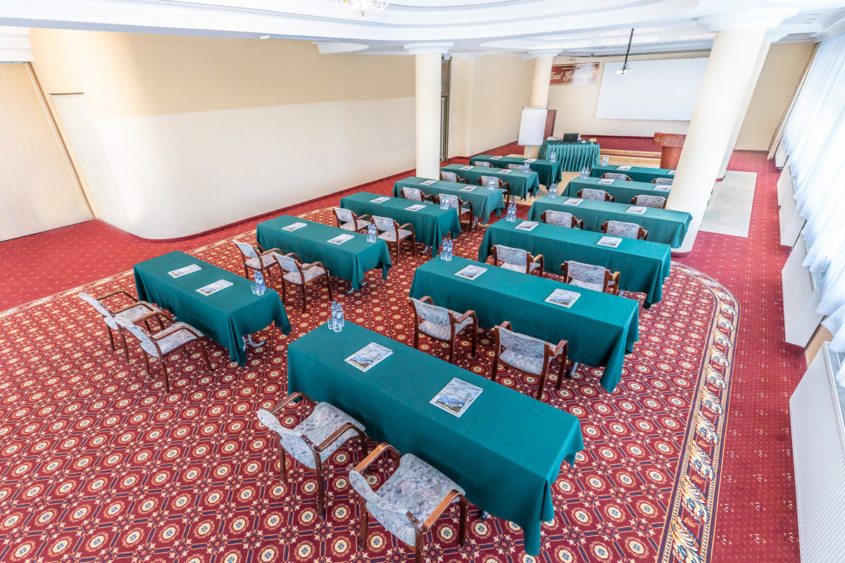 sala konferencyjna z zielonymi stołami i rzędami krzeseł oraz mównicą widoczną w tle
