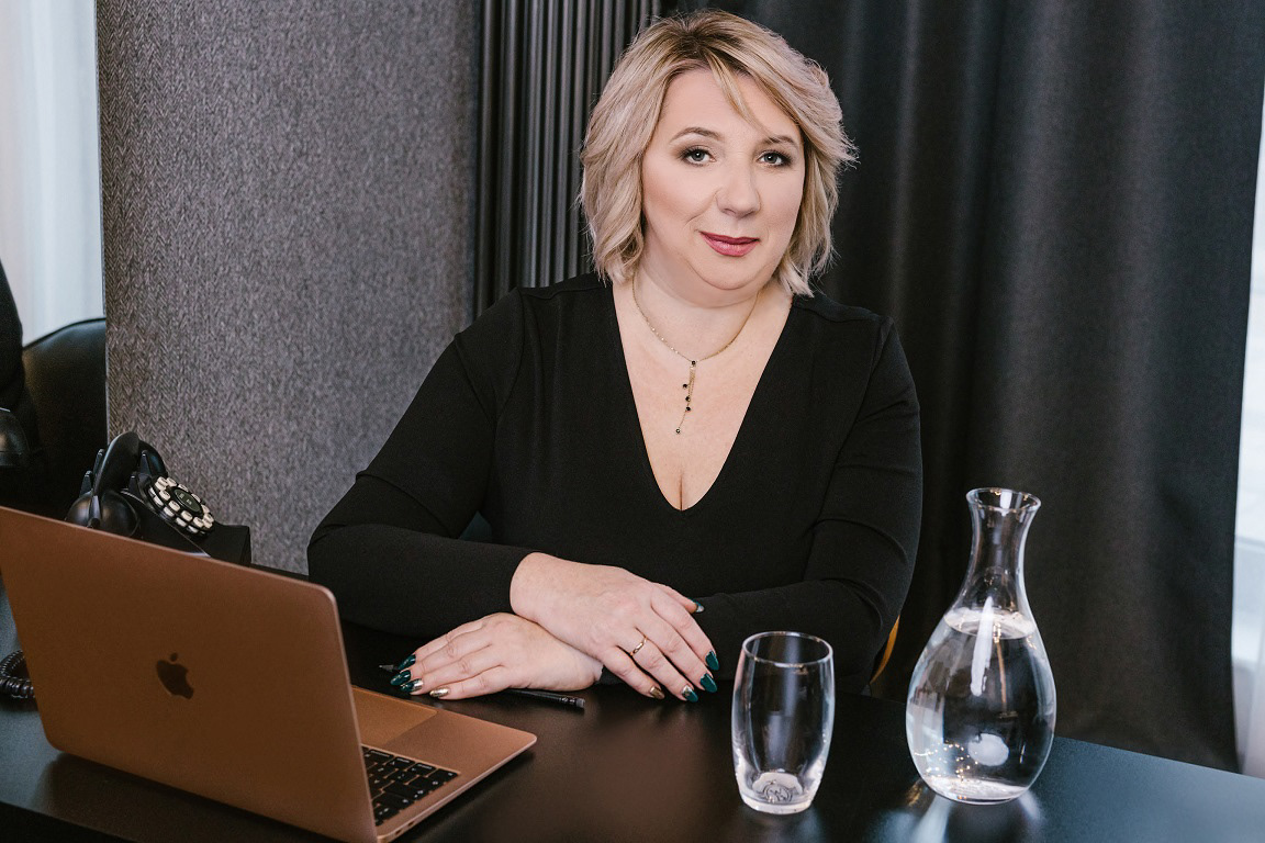Pani prezes Wodociągów Białostockich Beata Wiśniewska siedzi za biurkiem delikatnie uśmiechnięta a obok niej telefon, laptop i karafka z wodą
