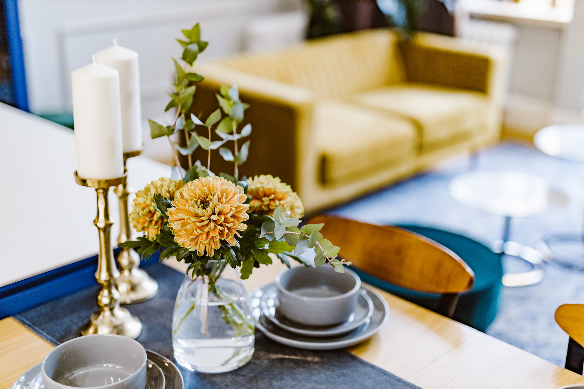Homestaging na jadalnianym stole z dwoma świecami w złotych świecznikach, kwatuszkami w wazoniku i żółtą sofą w tle