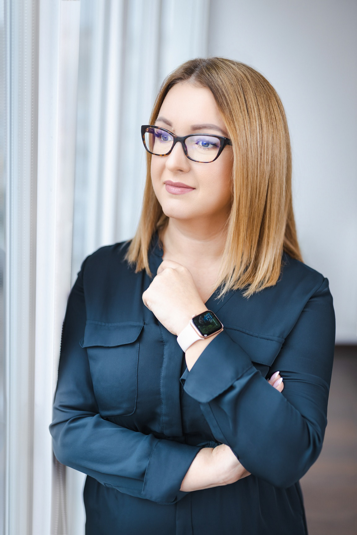 blondynka w okularach spogląda w okno, jest ładnie ubrana, ma smartwatch i jest zamyślona