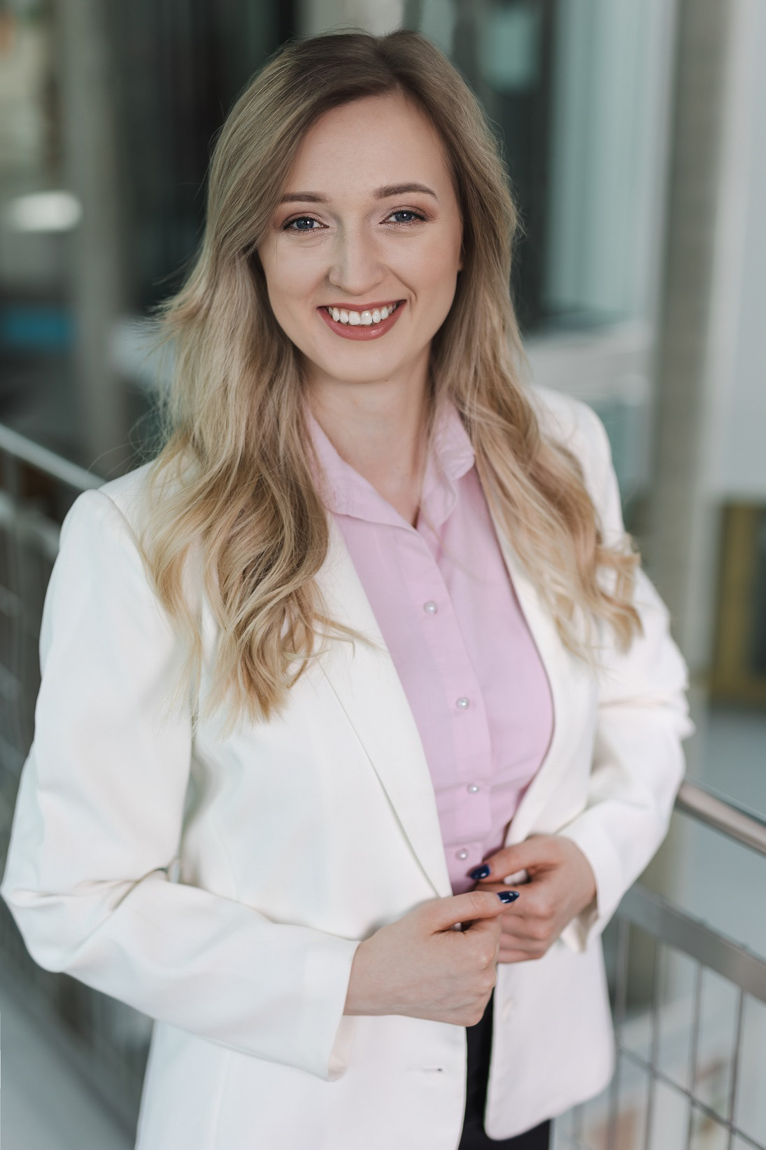Uśmiechnieta blondynka ubrana w różową koszulę i białą marynarkę opierająca się o stalową balustradę