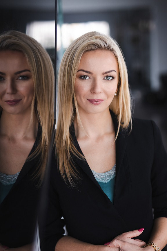uśmięchnieta pani stoi przy telewizorze, gdzie odbija się jej twarz. Patrzy prosto w obiektyw, ma blond włosy i ładny makijaż