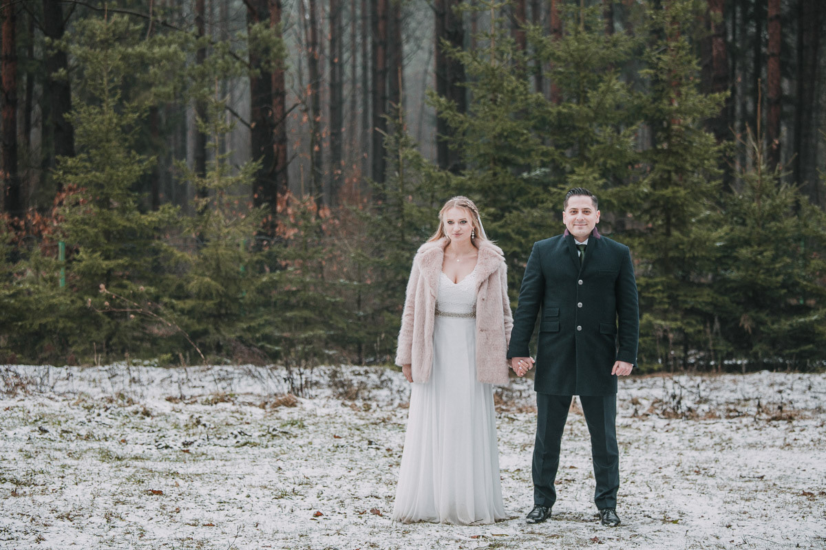 klasyczne zdjęcie pary młodej stojącej na śniegu na tle lasu, zimowa sesja ślubna pary młodej, fotografia ślubna Białystok