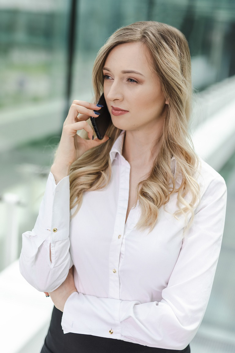 Blondynka z telefonem przy uchu, elegancko ubrana w białą koszulę z kręconymi włosami i szkłem w tle