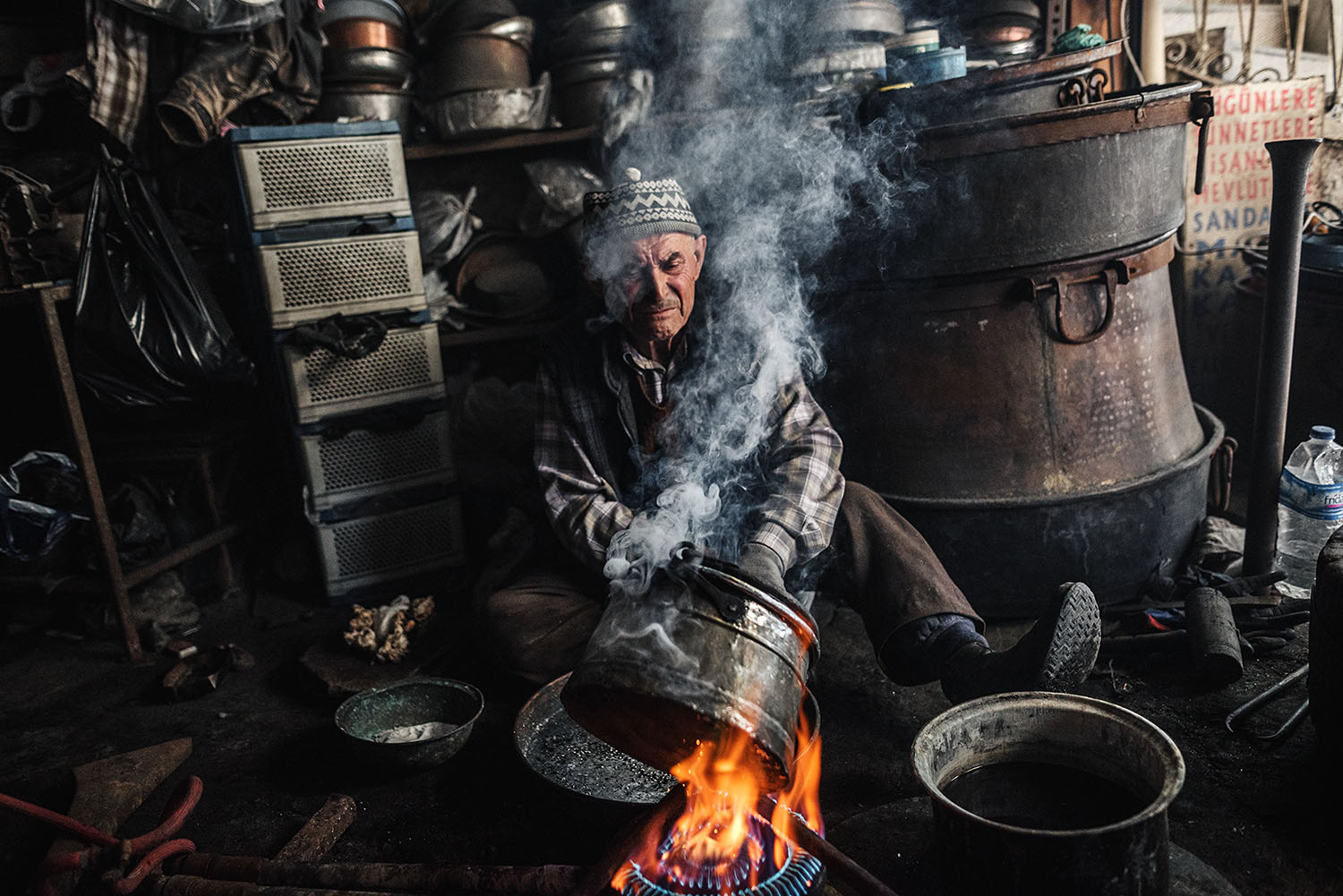 a tinsmith working on a large metal bowl over the fire.. There is visible smoke from the process of tinsmithing.