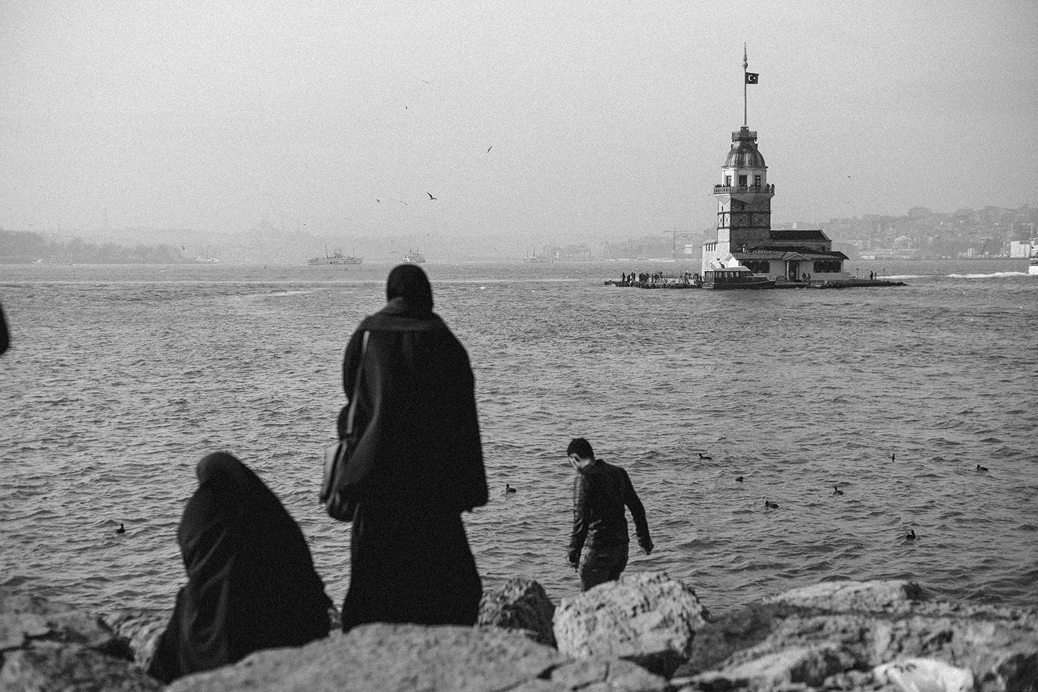 People sitting by the sea with the Maiden Tower in the background in a black and white image.
