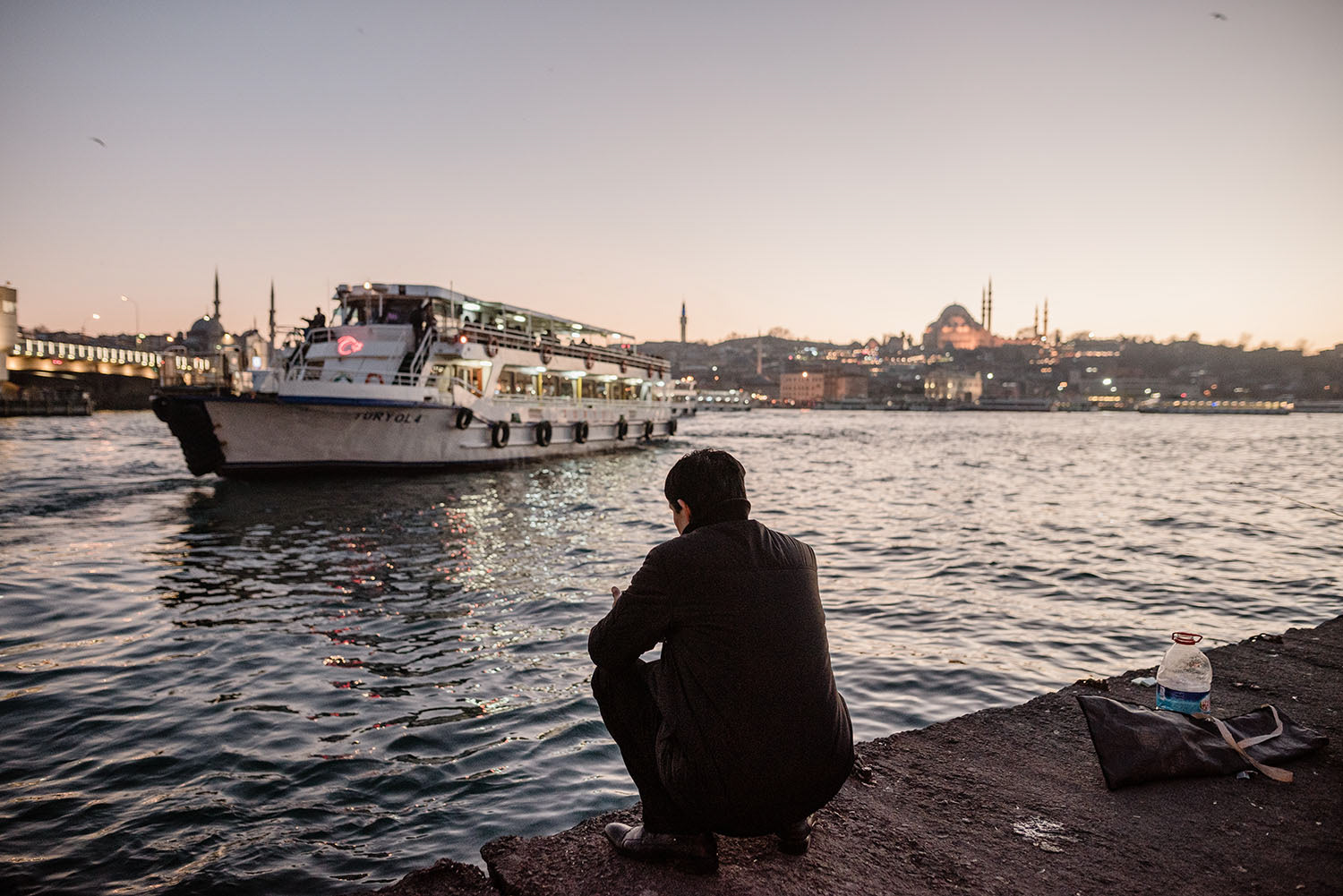 A man sitting by the sea in Haliç at sunset while a ferry is approaching the port in İstanbul, Türkiye.