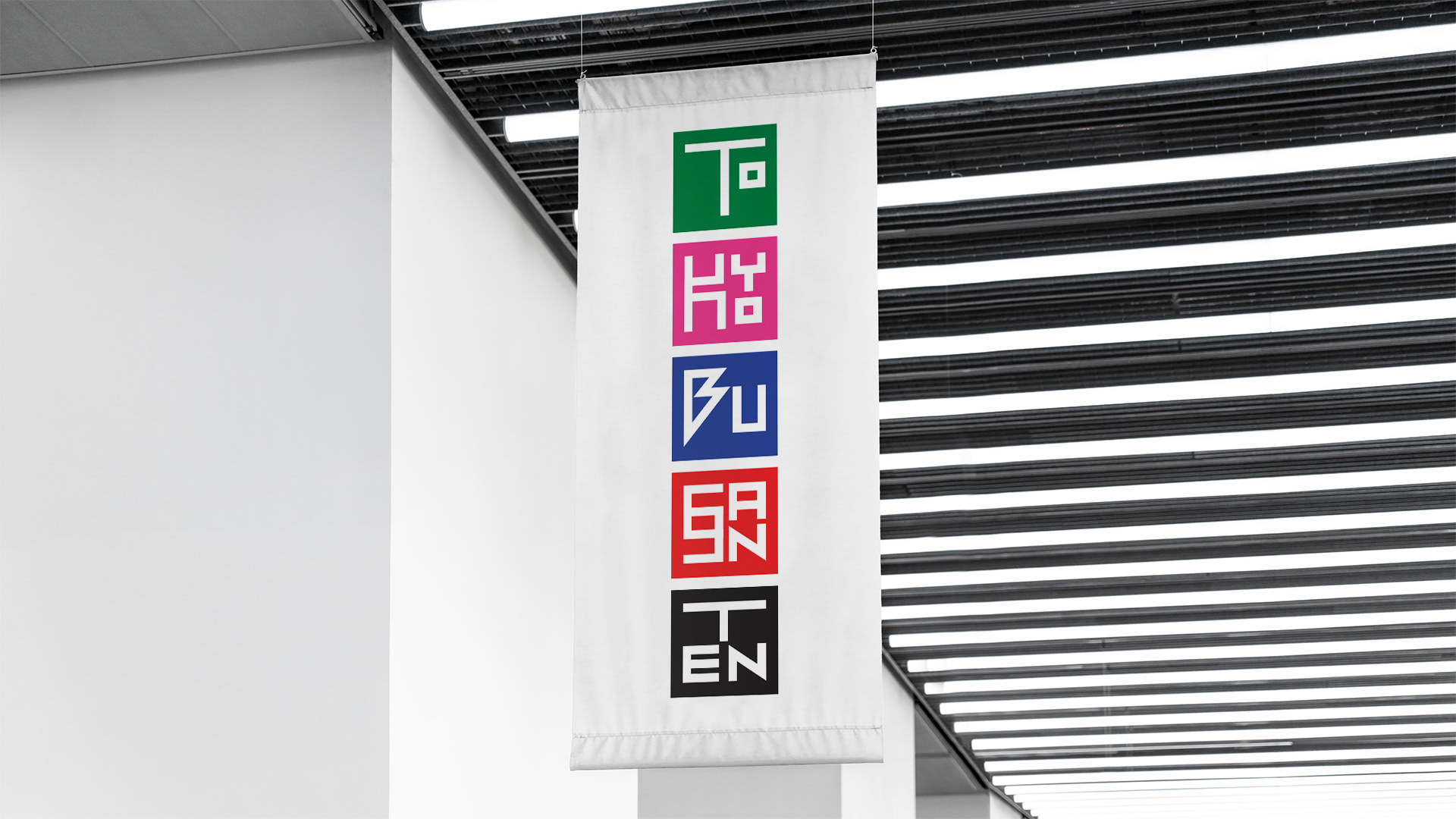 A vertical white exhibition banner hanging from a modern ceiling, showcasing a colorful stacked graphic logo that reads TOKYO BUSSANTEN in stylized, geometric block lettering.