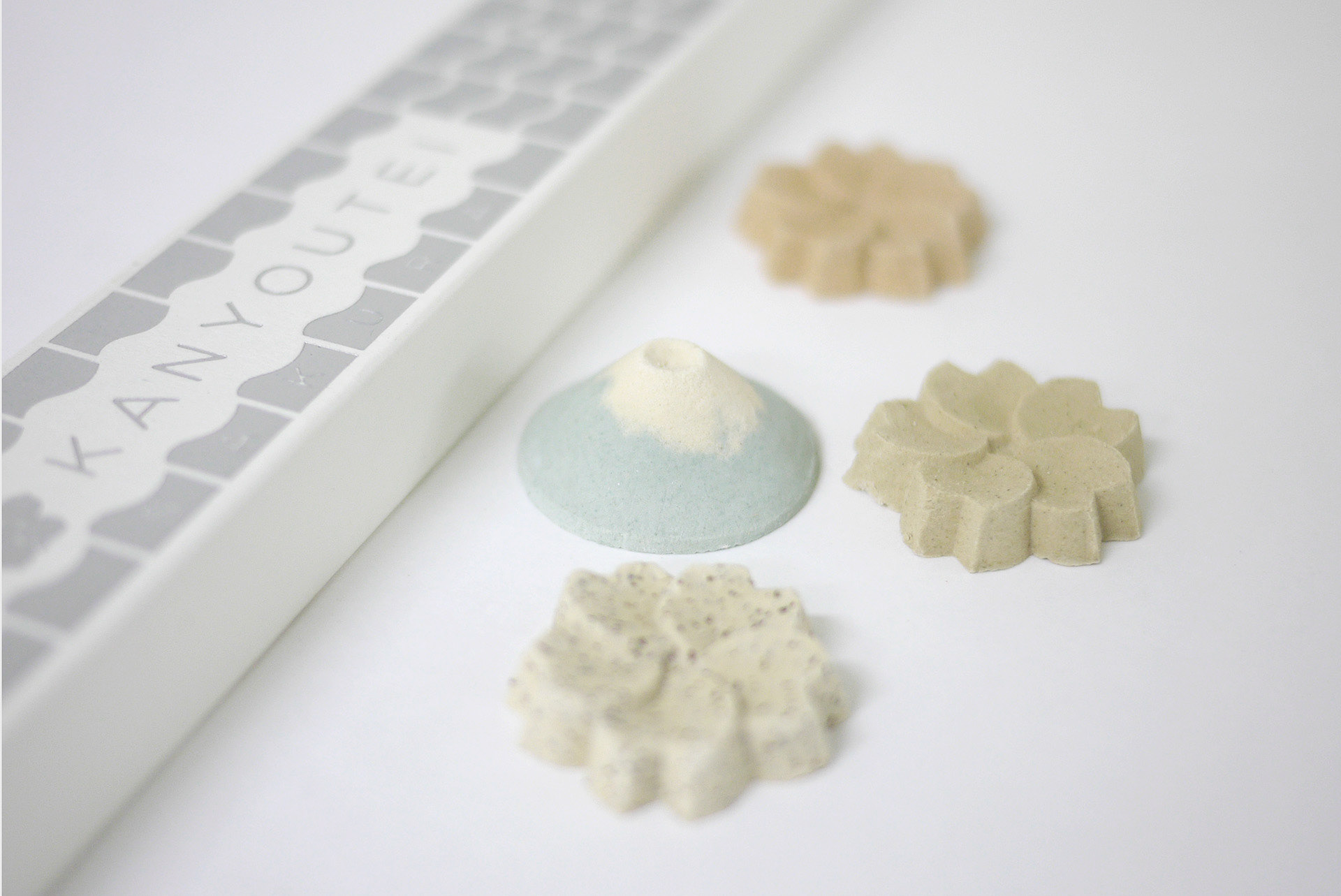 An elegant close-up shot of artisanal Japanese sugar confections (wasanbon) displayed next to their minimalist long white packaging. The packaging has the brand name "KANYOUTEI" printed vertically in a clean, modern serif font inside a grey scalloped pattern. Four distinct flower and mountain-shaped sweets are artfully scattered on a seamless white background. One confection is shaped like a light-blue Mount Fuji with a white snow-capped peak, while the other three are shaped like cherry blossoms in soft, natural earth tones of beige, pale green, and flecked ivory.