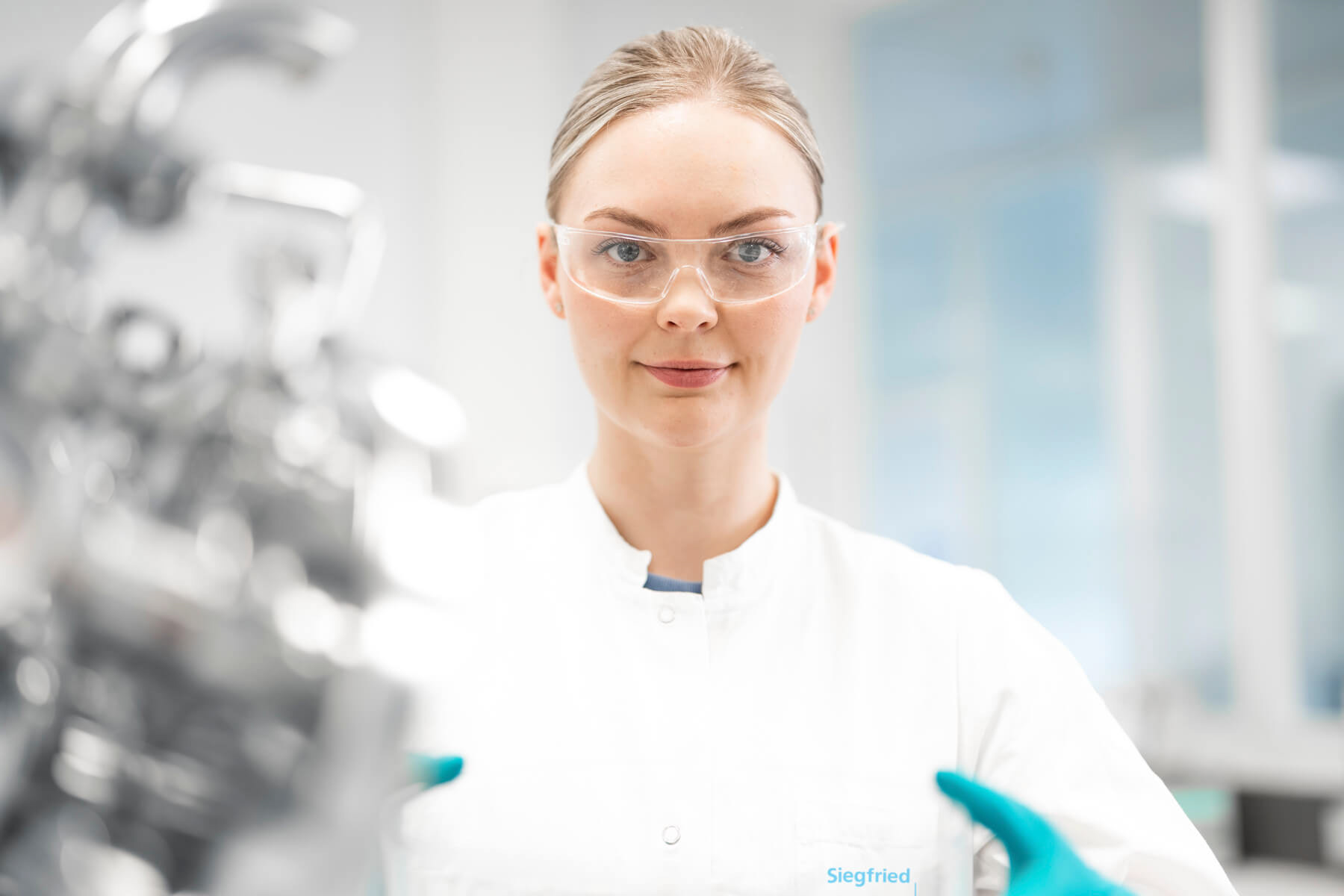 Labormitarbeiterin mit weißem Kittel und Schutzbrille schaut direkt in die Kamera. Edelstahl-Laborausrüstung unscharf im Vordergrund.
