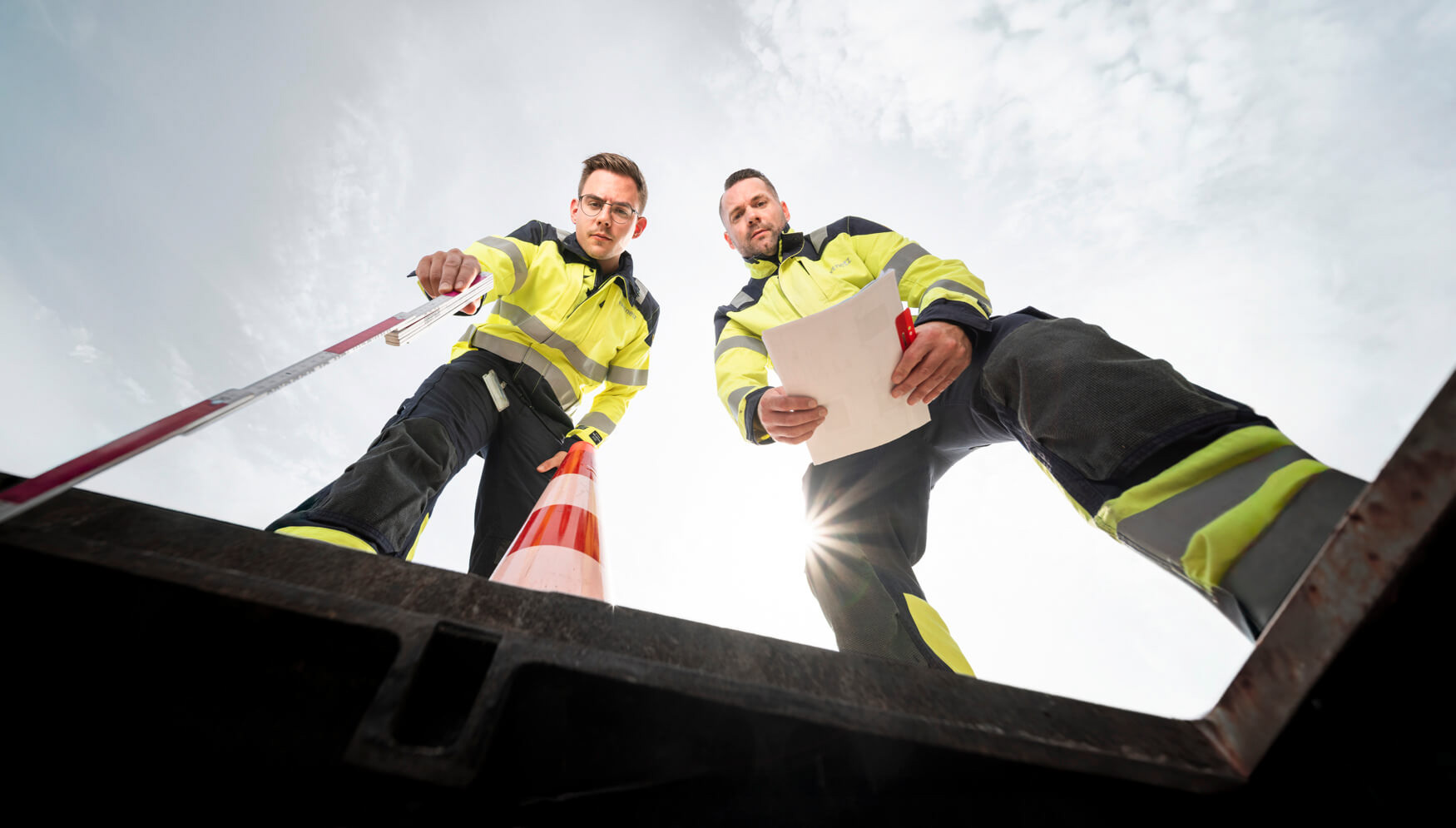 Zwei Mitarbeiter in Warnfarben schauen in einen Schacht, fotografiert in weitwinkliger Froschperspektive