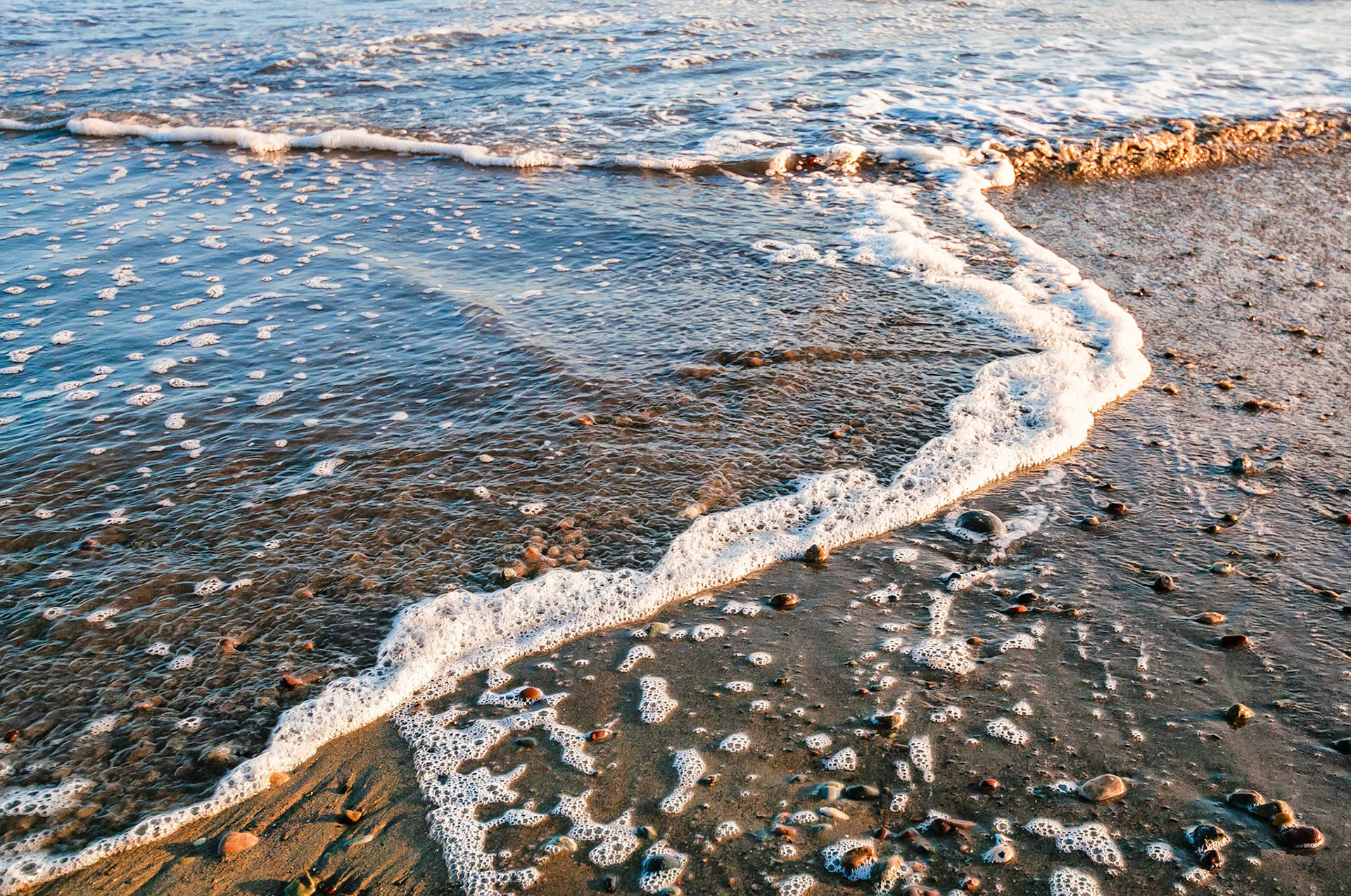 As waves expend their energy on a pebbly beach, they form dynamic patterns and curved lines of foam.
