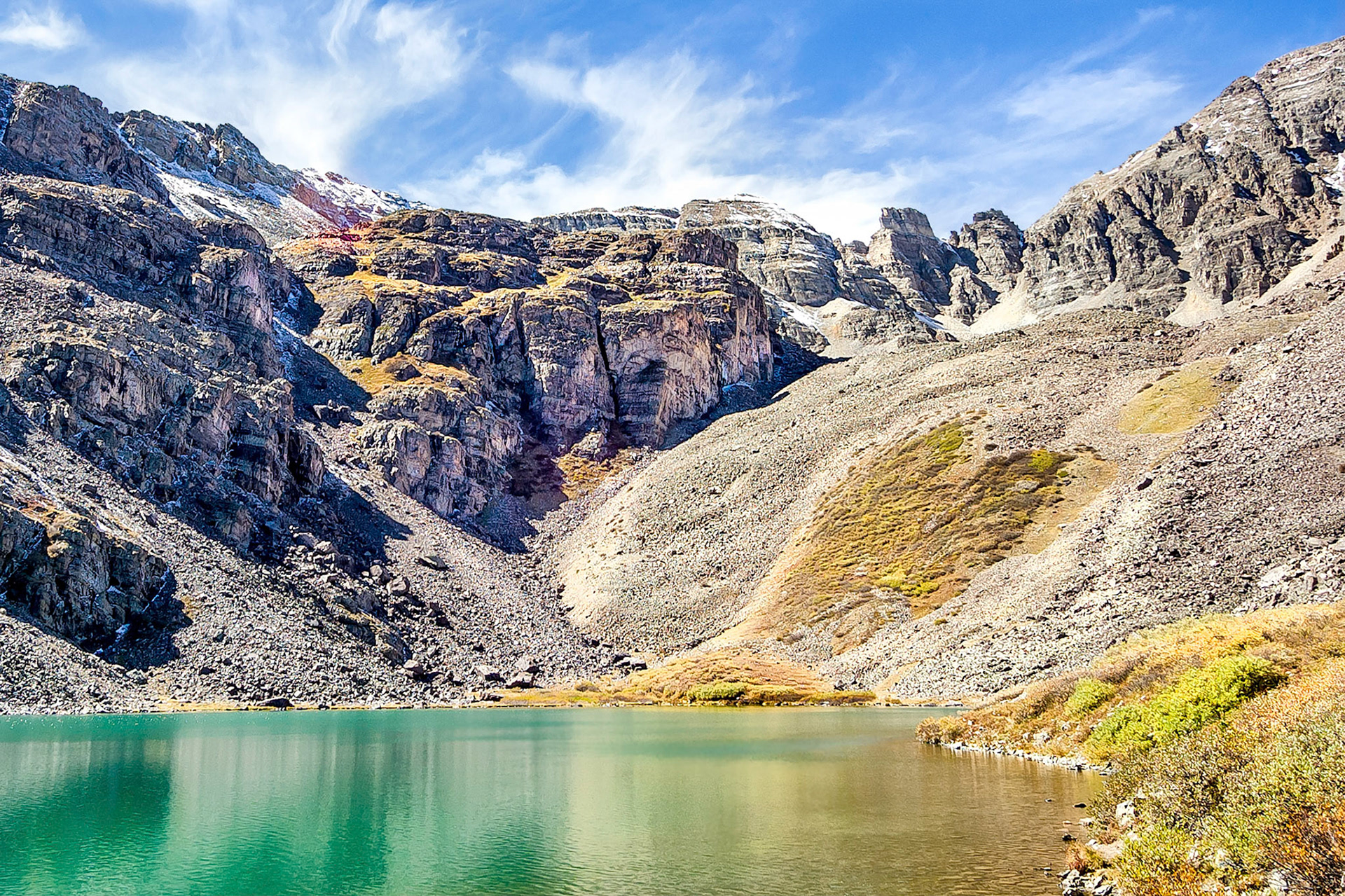 The clear waters of Cathedral Lake near Aspen, Colorado, USA, reflect rugged cliffs below a dramatic ridge line leading towards Cathedral Peak. (One can almost imagine two heart shapes in the image; one inverted heart in the partly cloudy sky above the ferruginous rock layers and yet another, formed by the scrub growing on the scree slope beyond the lake.)