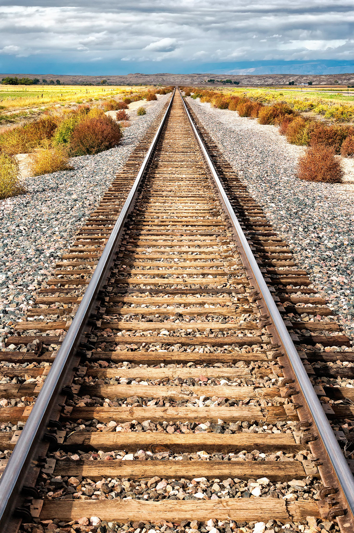 A railway line cuts across a seemingly endless plain in farming country near Worland, Wyoming, USA. As heavy clouds gather in the distance, crops growing beside the railway catch a patch of passing sunshine.
