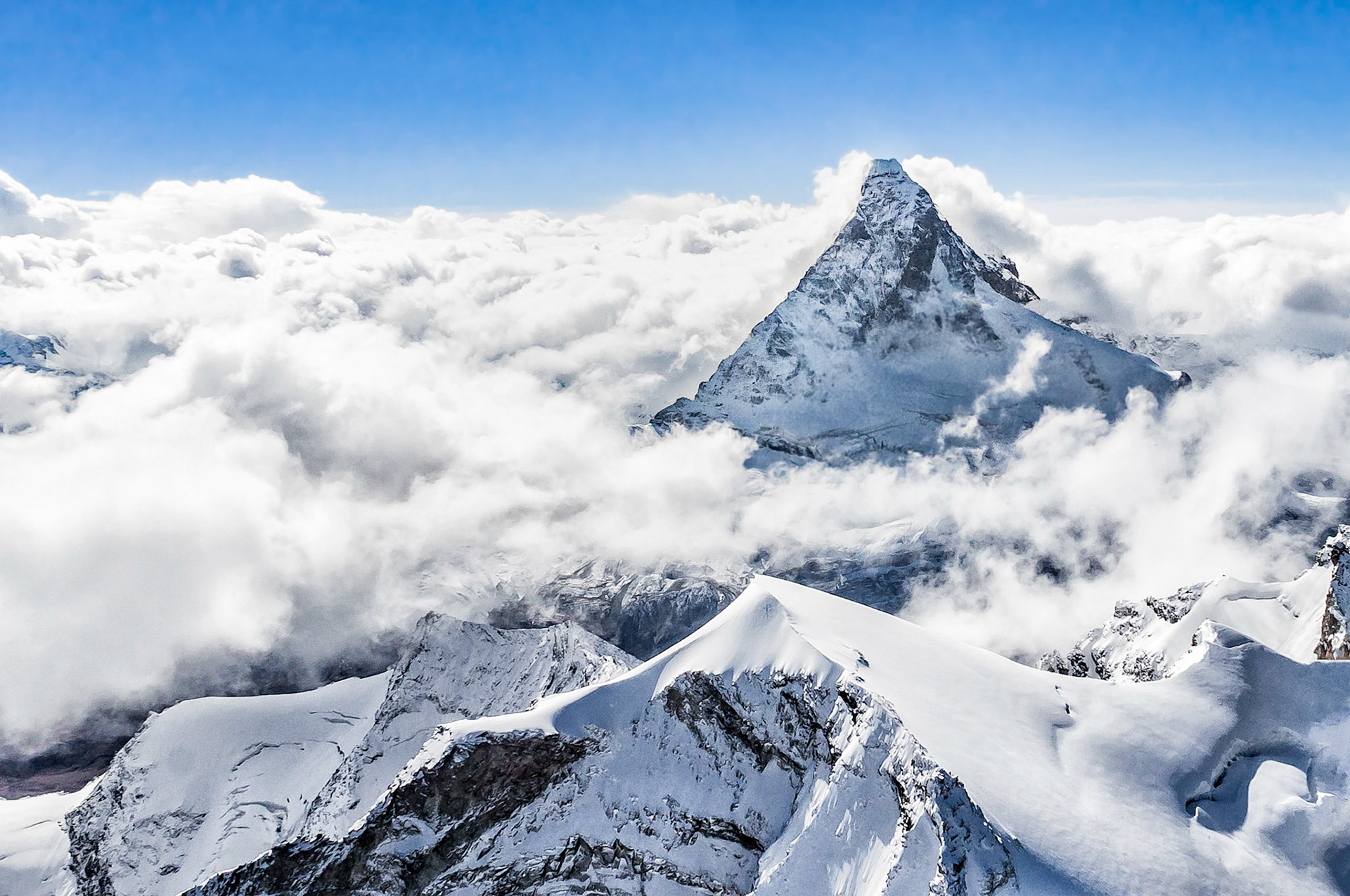 The iconic Matterhorn (4,478 m = 14,692 ft), located on the border between Switzerland and Italy, rises through a canopy of low hanging clouds into a clear blue sky above.