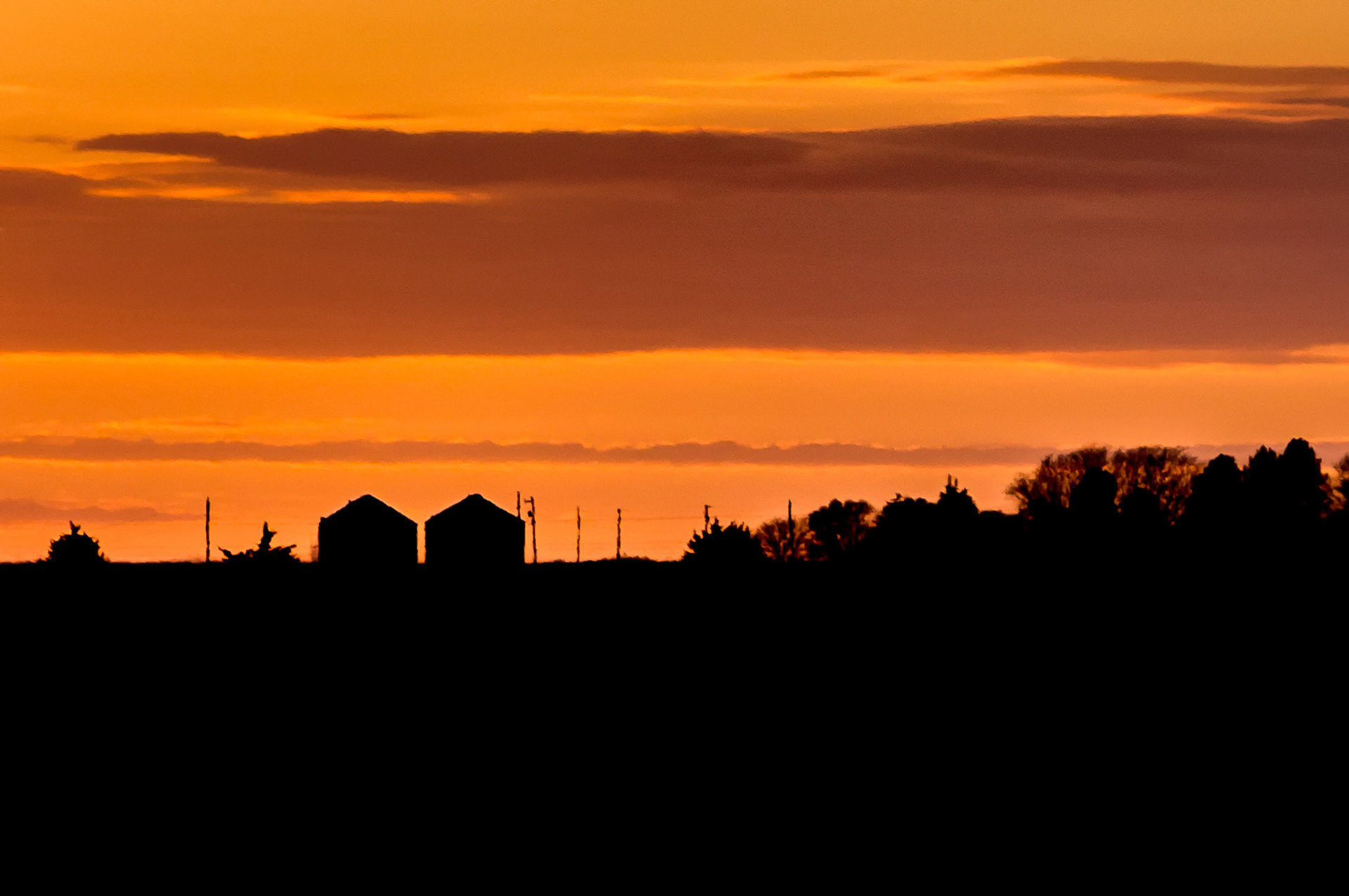 As the sun sinks below the horizon, grain storage silos stand in silhouette against a vivid evening sky near Paxton, Nebraska, USA.
