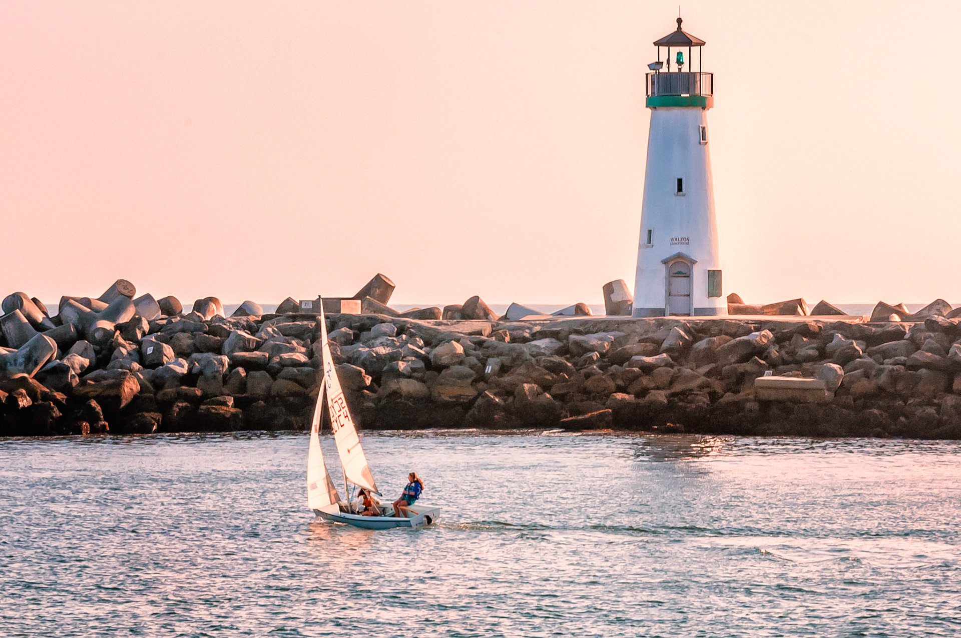The late afternoon sun casts a pinkish glow on the sky and waters near the Walton Lighthouse at Santa Cruz harbor in California, USA, as a light skiff sails past in a light breeze.