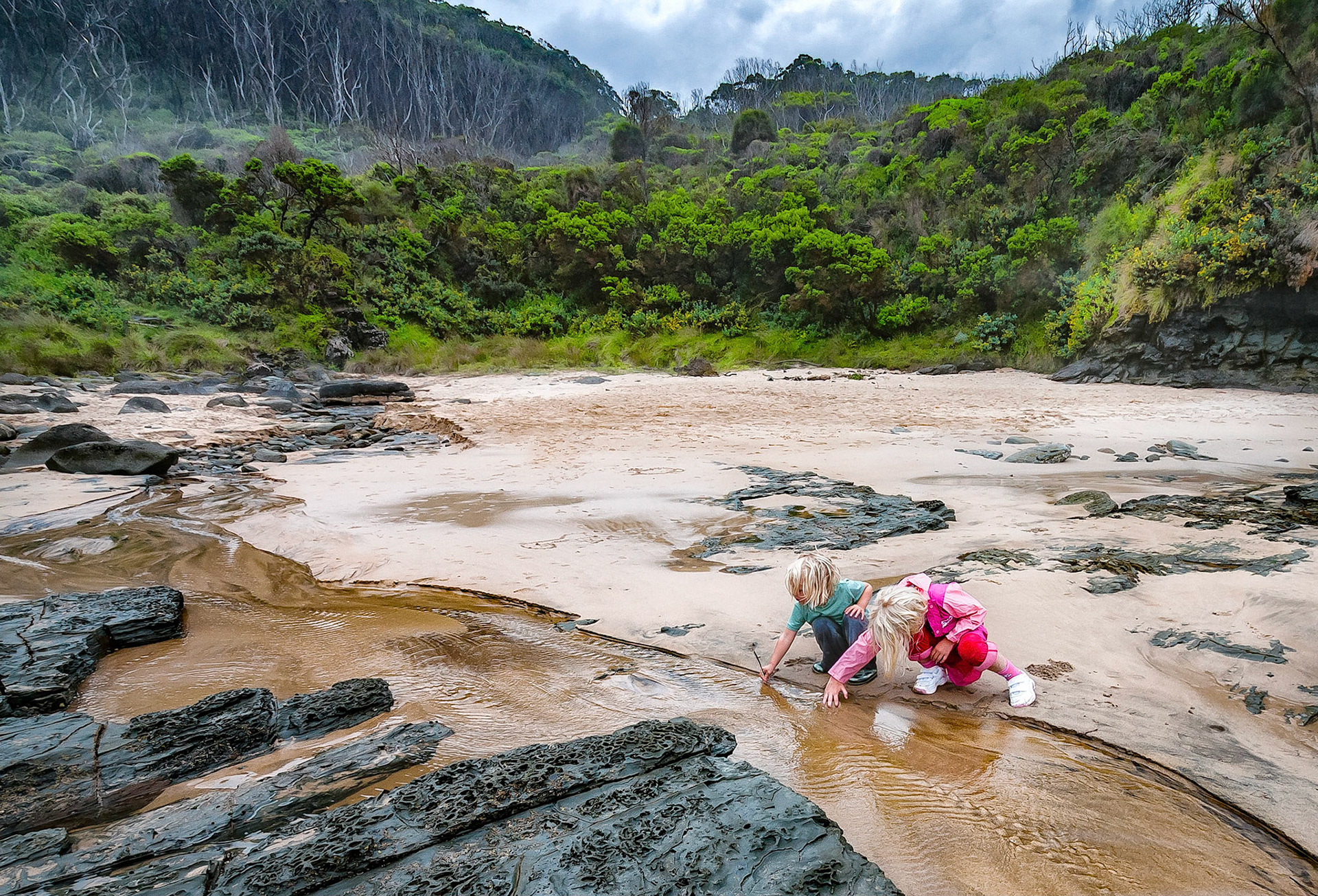 Children play in the water of a fresh water creek that flows across Shelley Beach on the Otway Coast, Victoria, Australia on a chilly, cloudy day.
