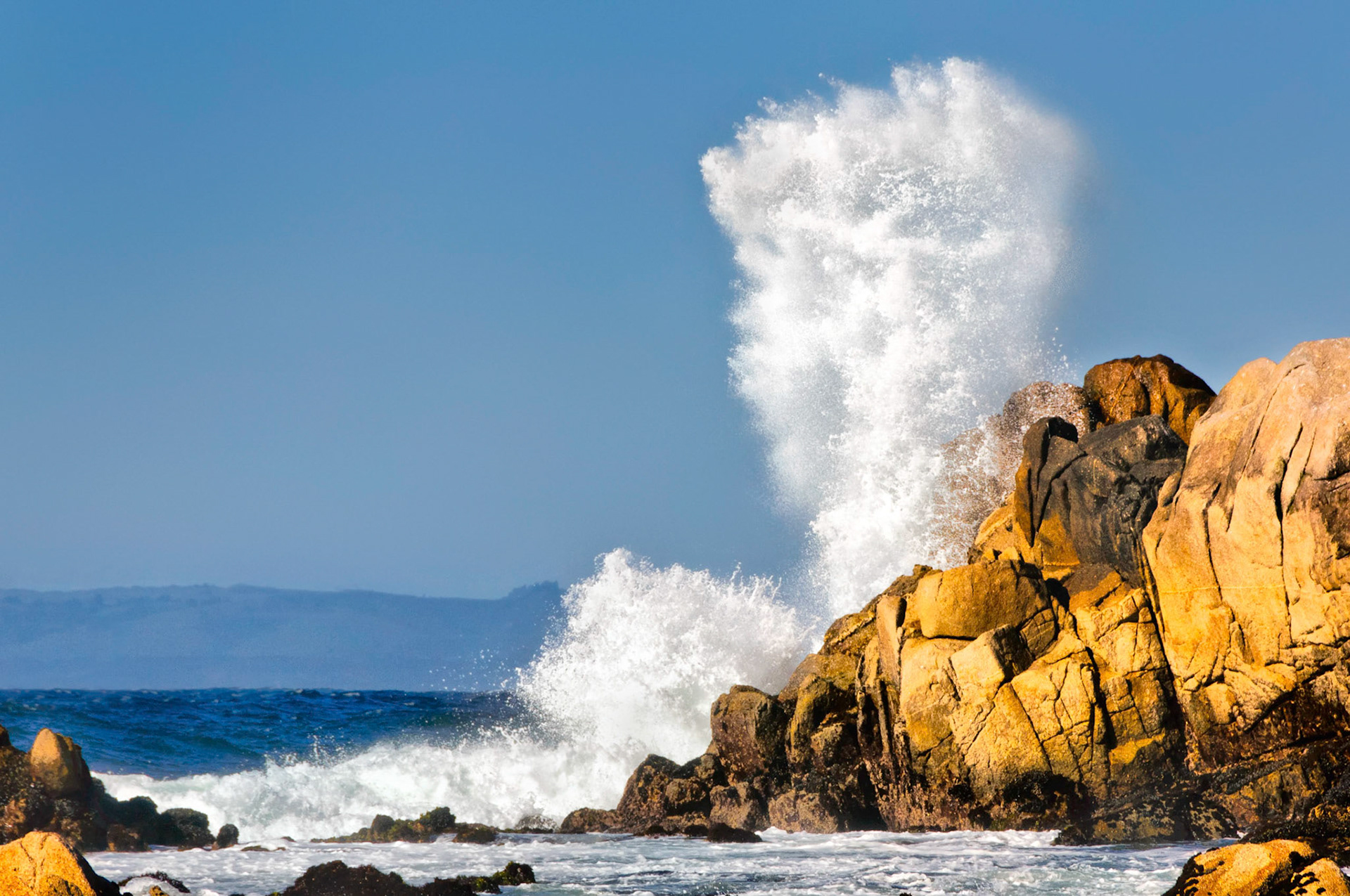 Rocks glow in the late afternoon sun as it lights up the powerful waves crashing against a weathered headland along the Monterey Bay coastline; California, USA.