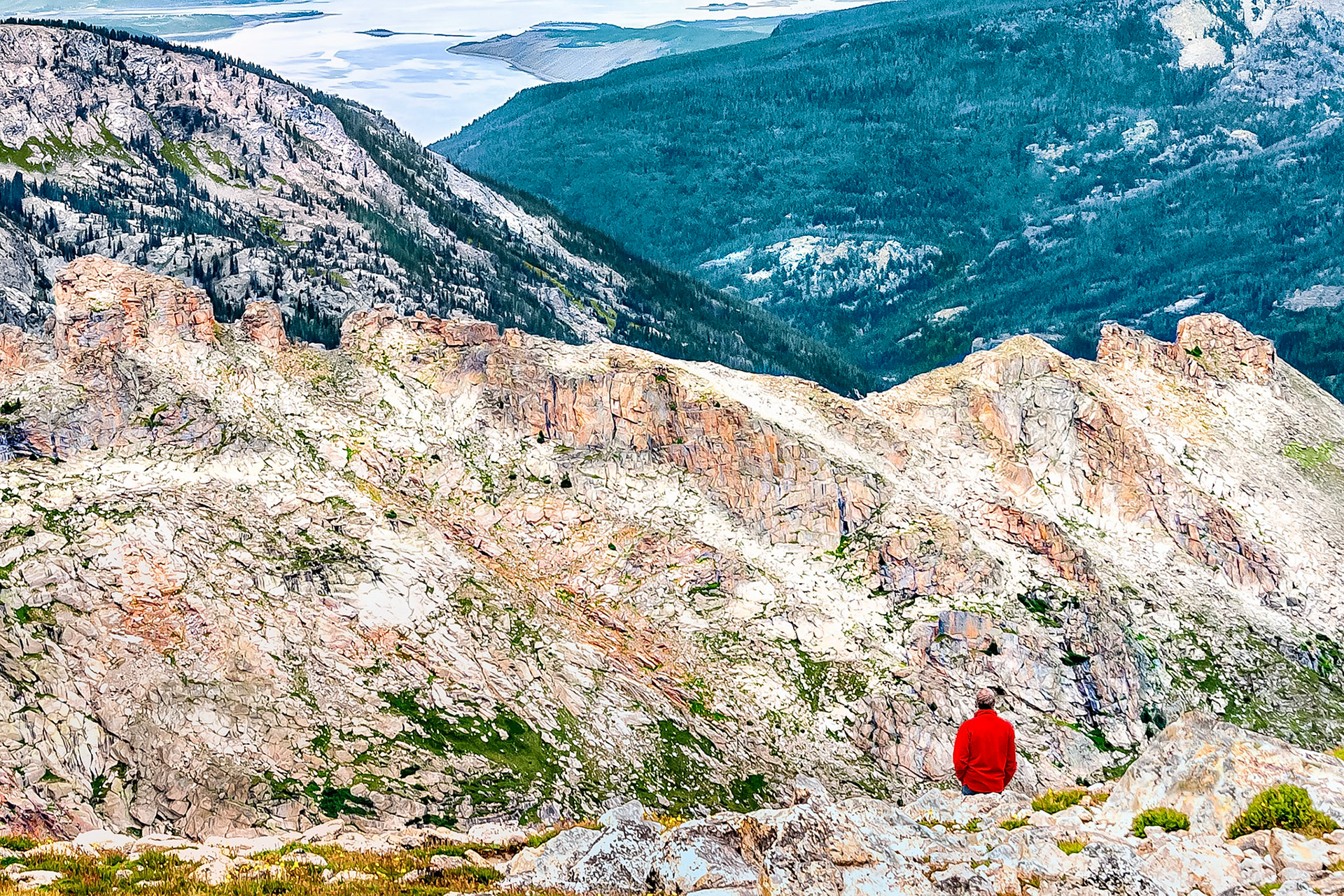 A hiker takes in the views from Pawnee Pass, (12,542 ft = 3,823 m), on the Rocky Mountain Continental Divide in the Indian Peaks Wilderness Area. Grand Lake, the largest natural lake in Colorado, USA, (at 8,369 feet = 2,551 m), shimmers in the distance under cloudy skies.
