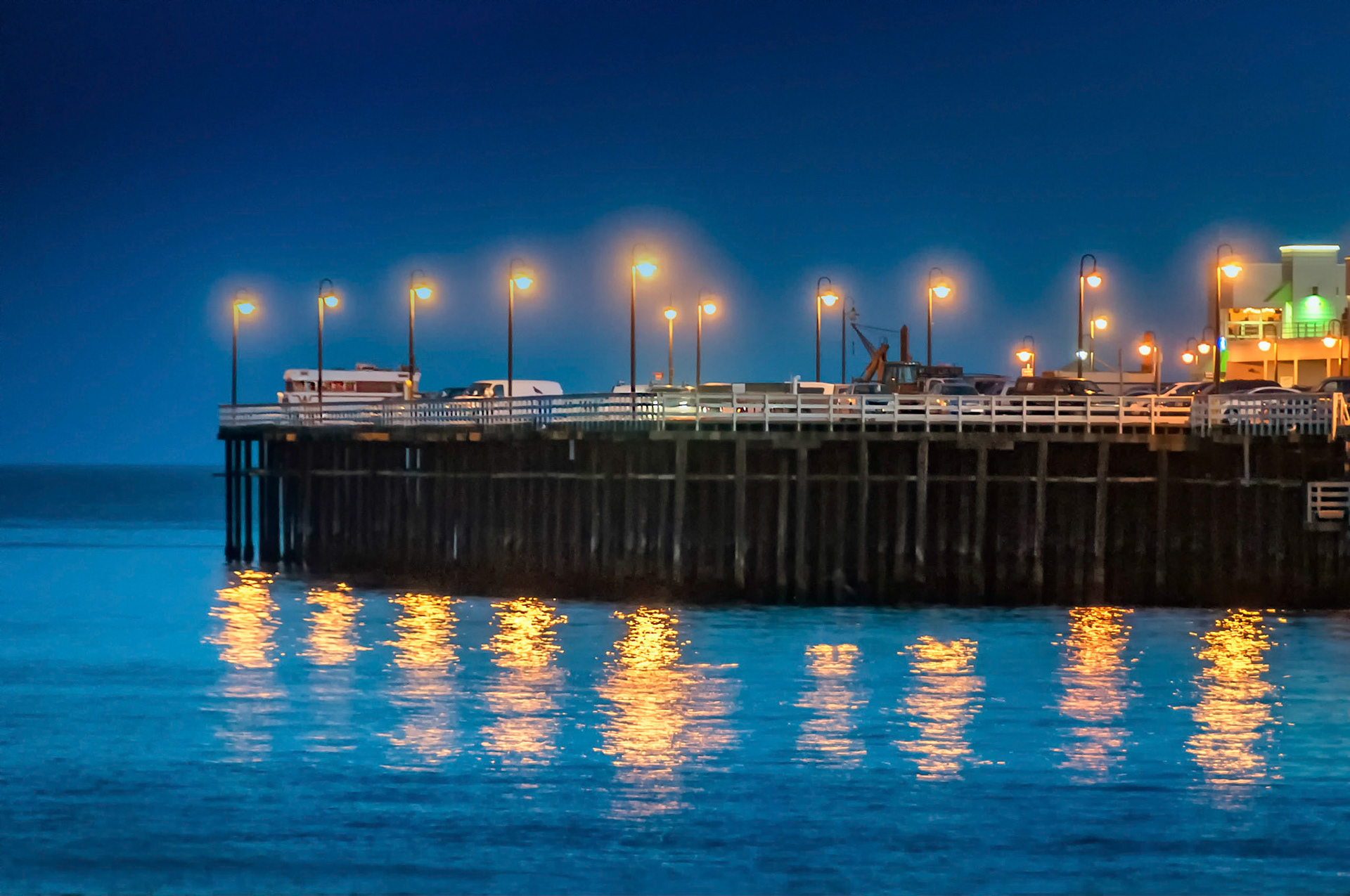 As night falls over Monterey Bay at Santa Cruz, California, a light sea mist creates glowing halos around the street lamps on the deck of the jetty. The lights form their shimmering reflections on the calm waters.