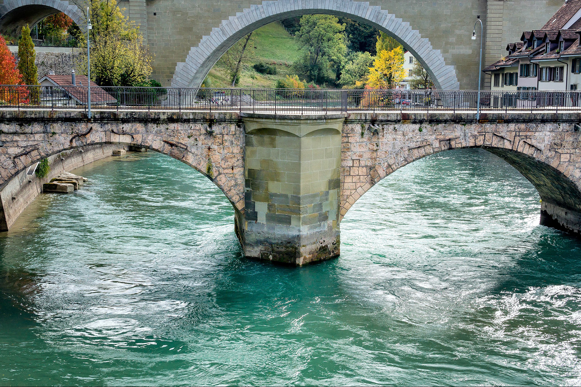Reaching across the River Aare, "The Untertorbrücke" is the oldest bridge in Bern, Switzerland. Completed in 1489, this granite clad, sandstone core structure replaced an earlier wooden bridge that served from 1256. In the background is "The Nydeggbrücke", (1840),  featuring the widest stone arch in Europe. Part of the famous Bern Bear Park can be seen through this span.