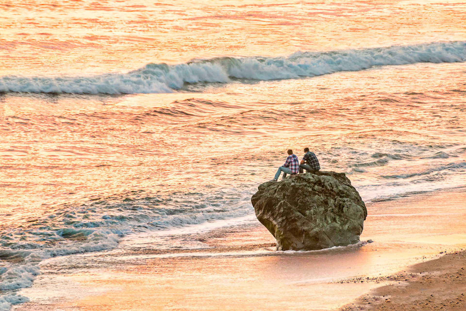 Two friends seated on a weathered rock on the beach, enjoy some quiet time while watching the sunset near San Simeon, California, USA.