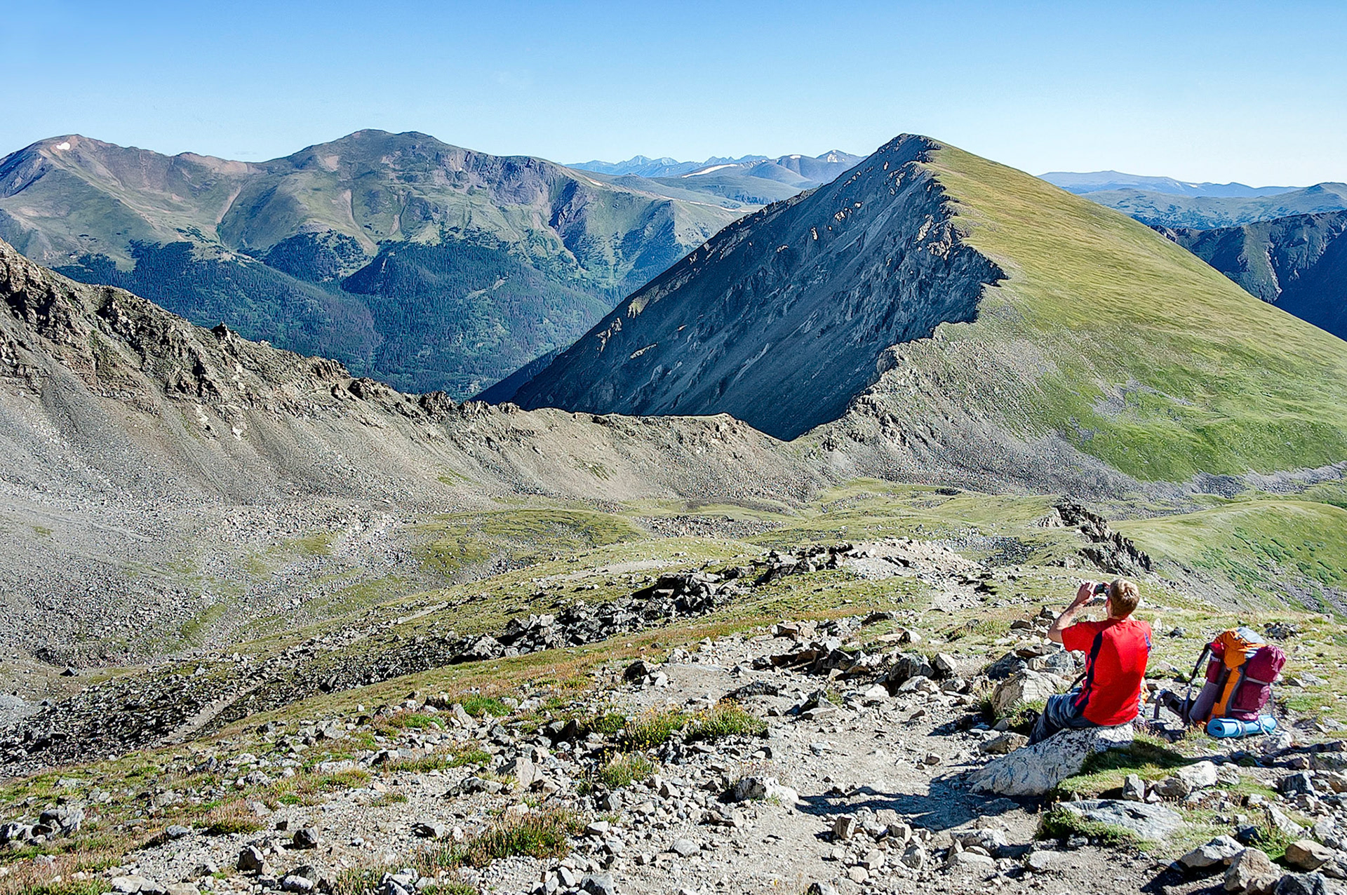 A hiker rests on the scree slopes of Grey's Peak, Colorado, USA and snaps a photograph of the Rocky Mountain terrain.
