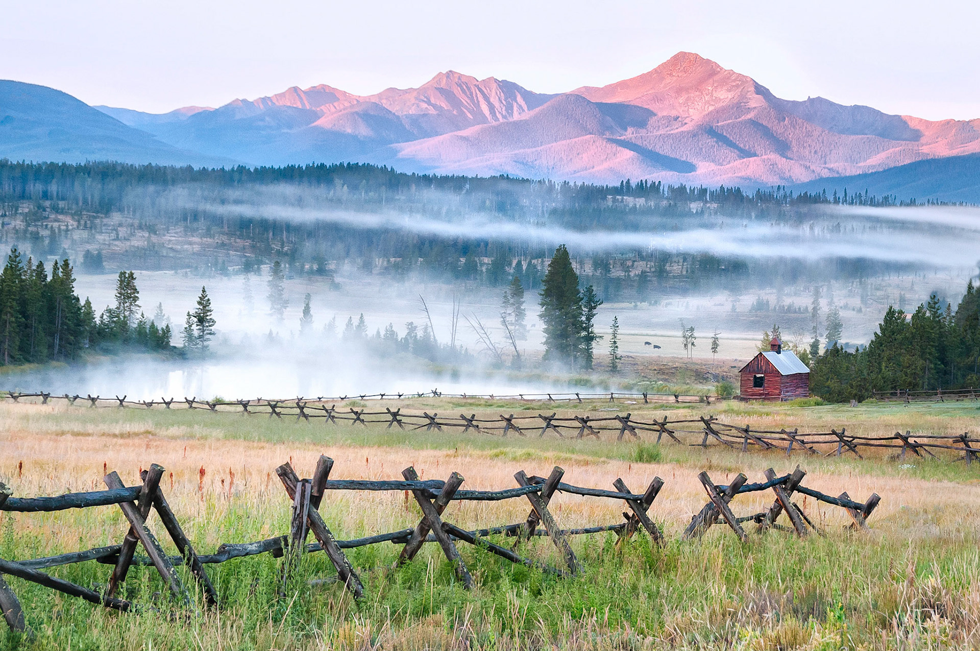 Sunlight clips Byers Peak near Fraser, Colorado, USA as the early morning fog lifts from the valley floor and mists rise from a water catchment pond. An old unused barn and dilapidated log-rail fences yield to the ravages of time.