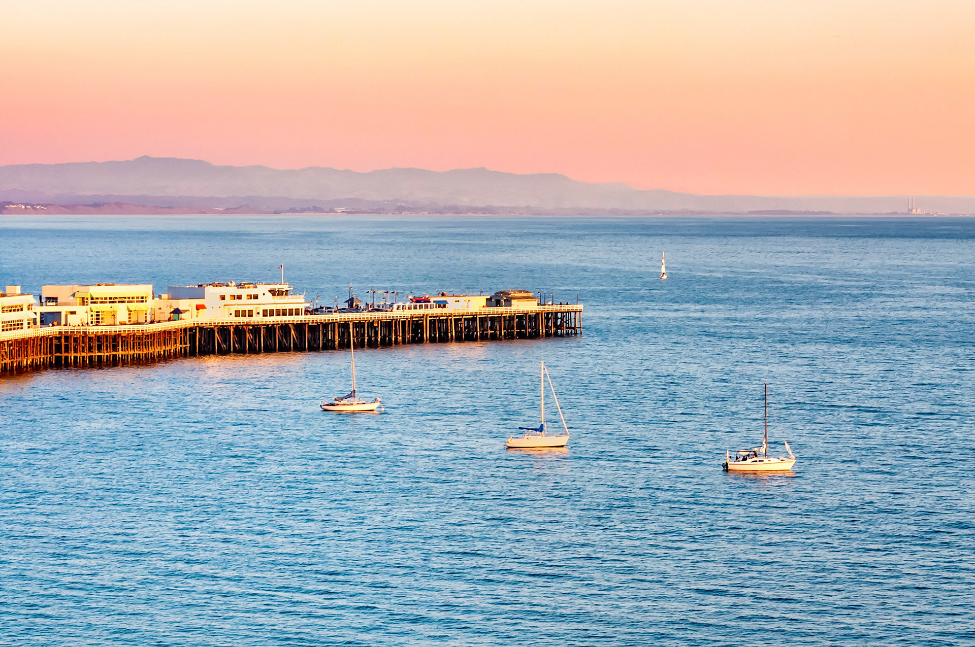 As evening falls over Monterey Bay, California, a warm orange glow develops in the sky. Reflections of the setting sun from the Santa Cruz jetty and the anchored sailing boats, appear in the peaceful waters.