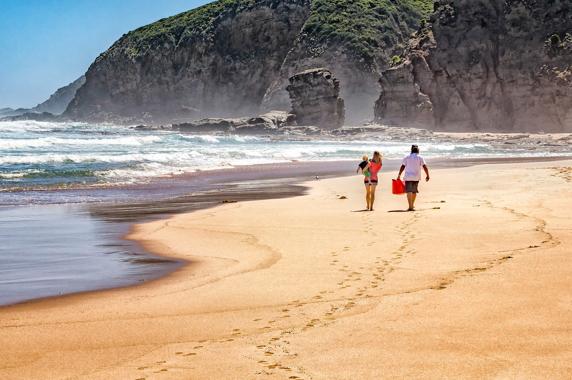 A young family strolls along a deserted beach towards Castle Cove in the Great Otway National Park near Apollo Bay, Victoria, Australia. Castle Rock, shrouded in a light sea mist, stands defiant against the erosive forces of the wind and relentless surf of Bass Strait.
