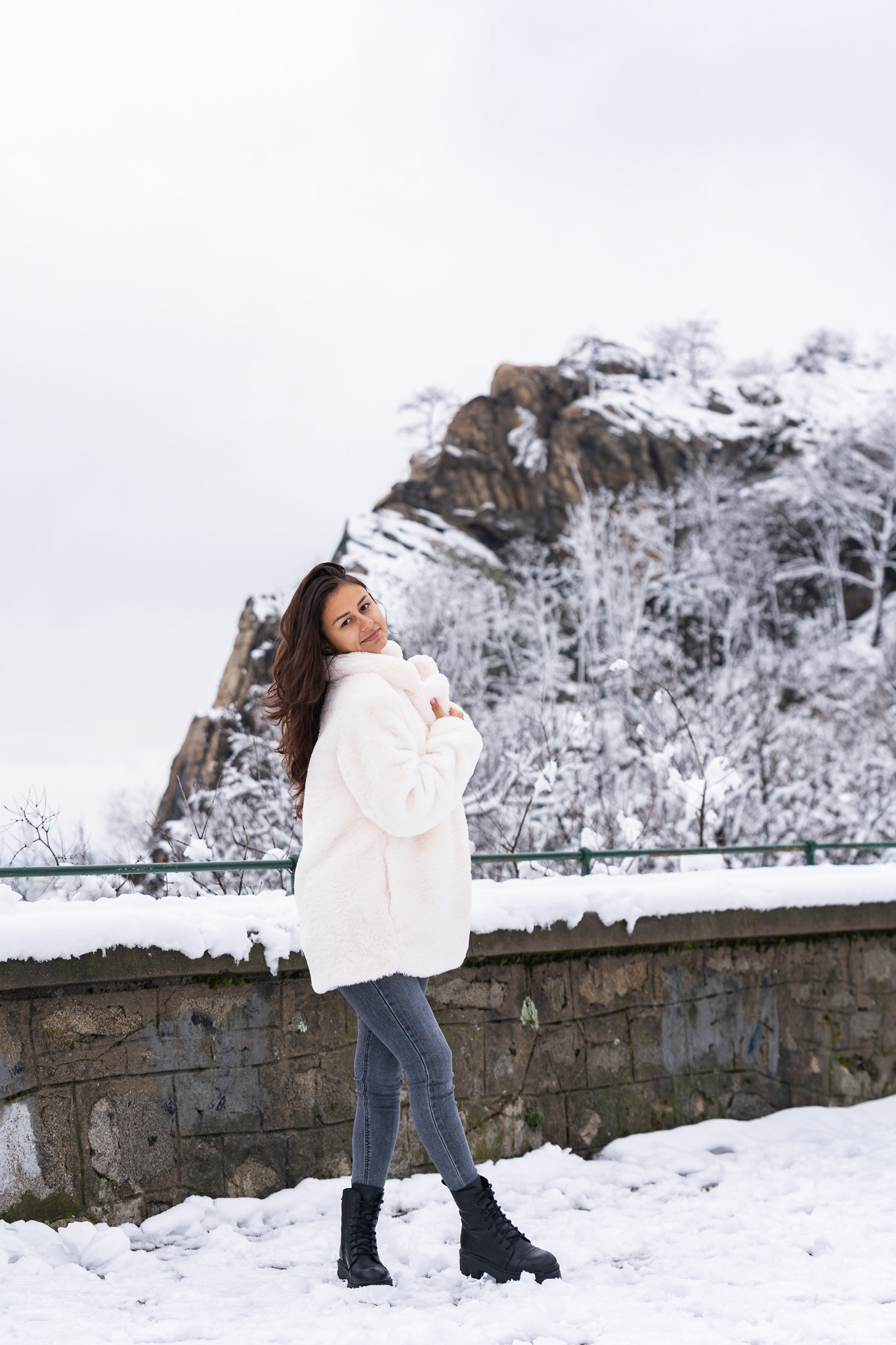 Fashion portrait of a brunette model in a white fur coat standing against a dramatic snow-covered mountain backdrop. Outdoor editorial photography by Arcane Media, UK.
