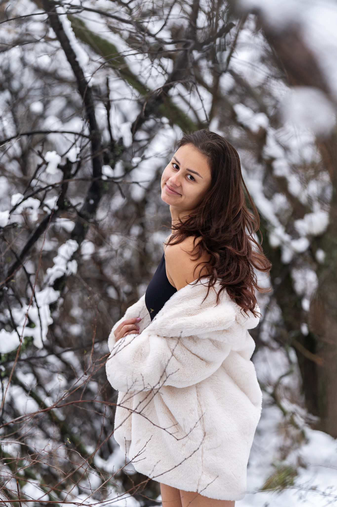 Editorial winter portrait of a curly haired female model in a white coat posing playfully in a snowy landscape. Natural light photography by Tenyo Barbolski.